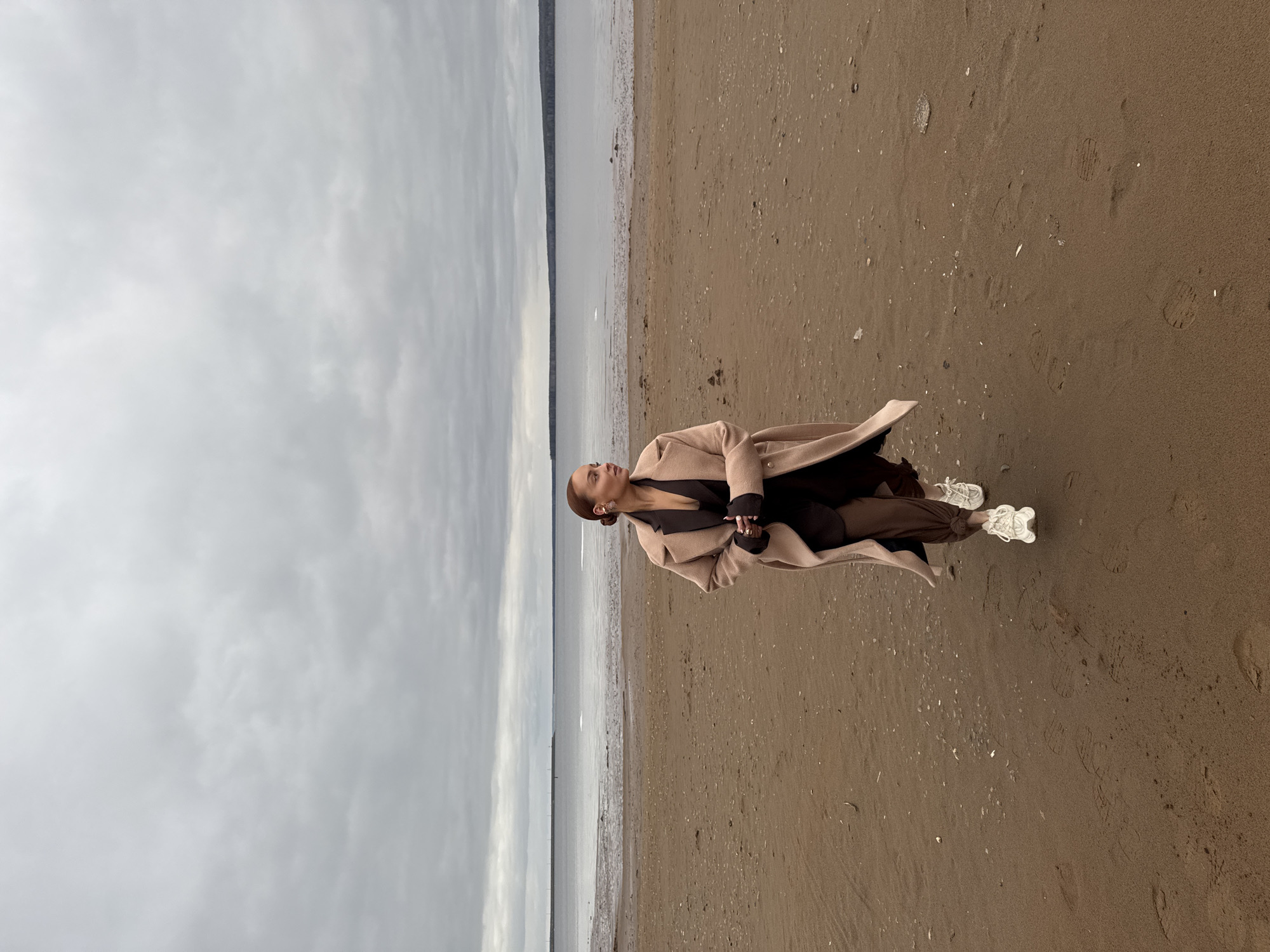 A full, sideways photograph shows a light-skinned woman, wearing brown pants, black top and a light tan overcoat, on a gray, sandy beach in Chaikovsky, Russia. She wears white sneakers, and her hair is pulled back. Her head is turned toward the body of water to her left, which separates the beach from a strip of tree-covered land. The sky above the water is gray and cloudy. The sand is flat. There are visible footprints in the sand.