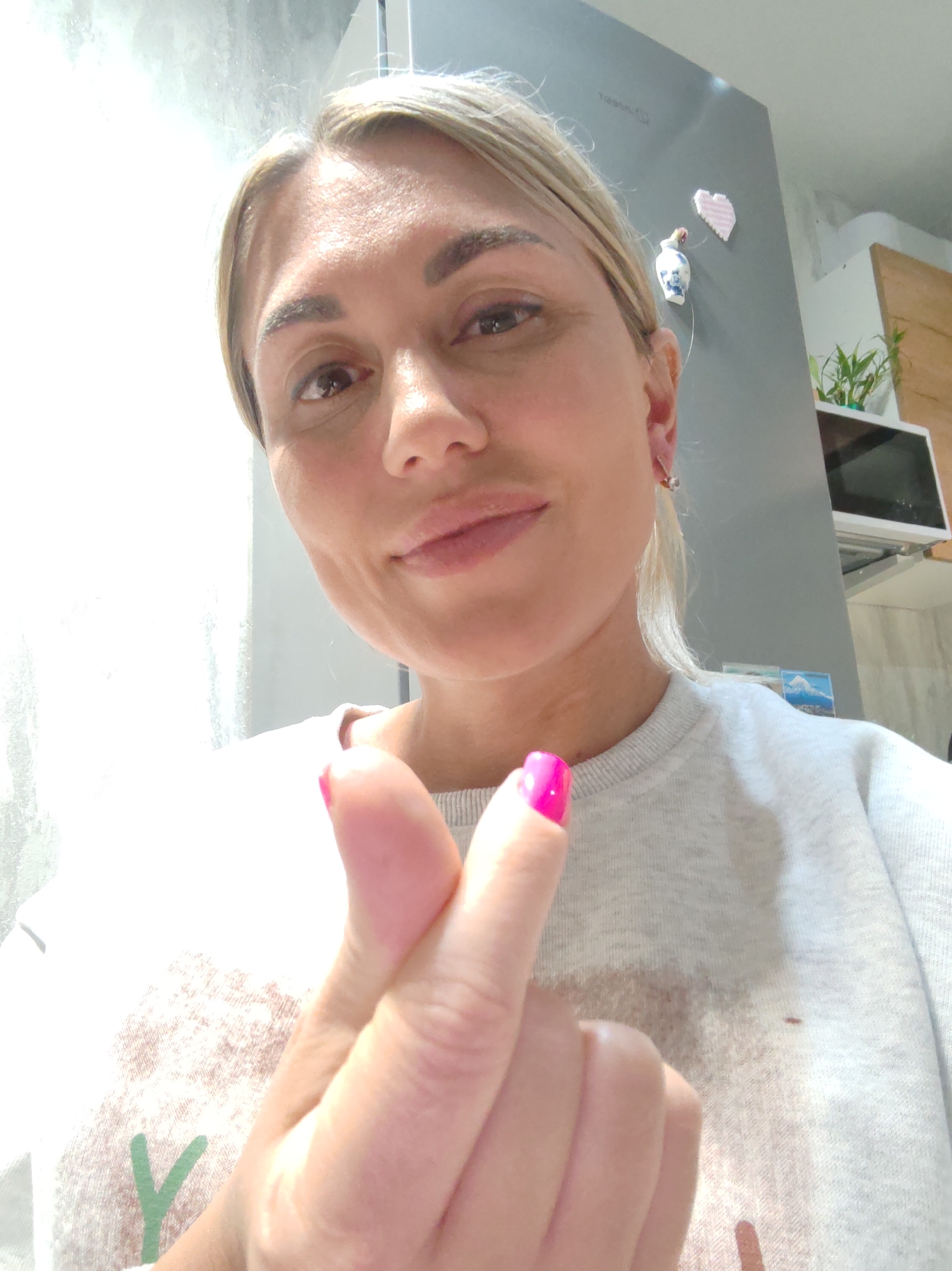 A selfie of a woman with blonde hair is taken in a kitchen. The woman is in the foreground, making a finger heart gesture. She is wearing a gray sweatshirt. In the background, a refrigerator is visible, adorned with a pink heart magnet and a small decorative vase. A potted plant and a microwave are also visible on a shelf above the refrigerator. The lighting is bright, suggesting an indoor environment. The presence of a refrigerator and kitchen appliances indicates a domestic setting, common in residential areas. The woman's gesture and cheerful expression suggest a casual and friendly interaction.
