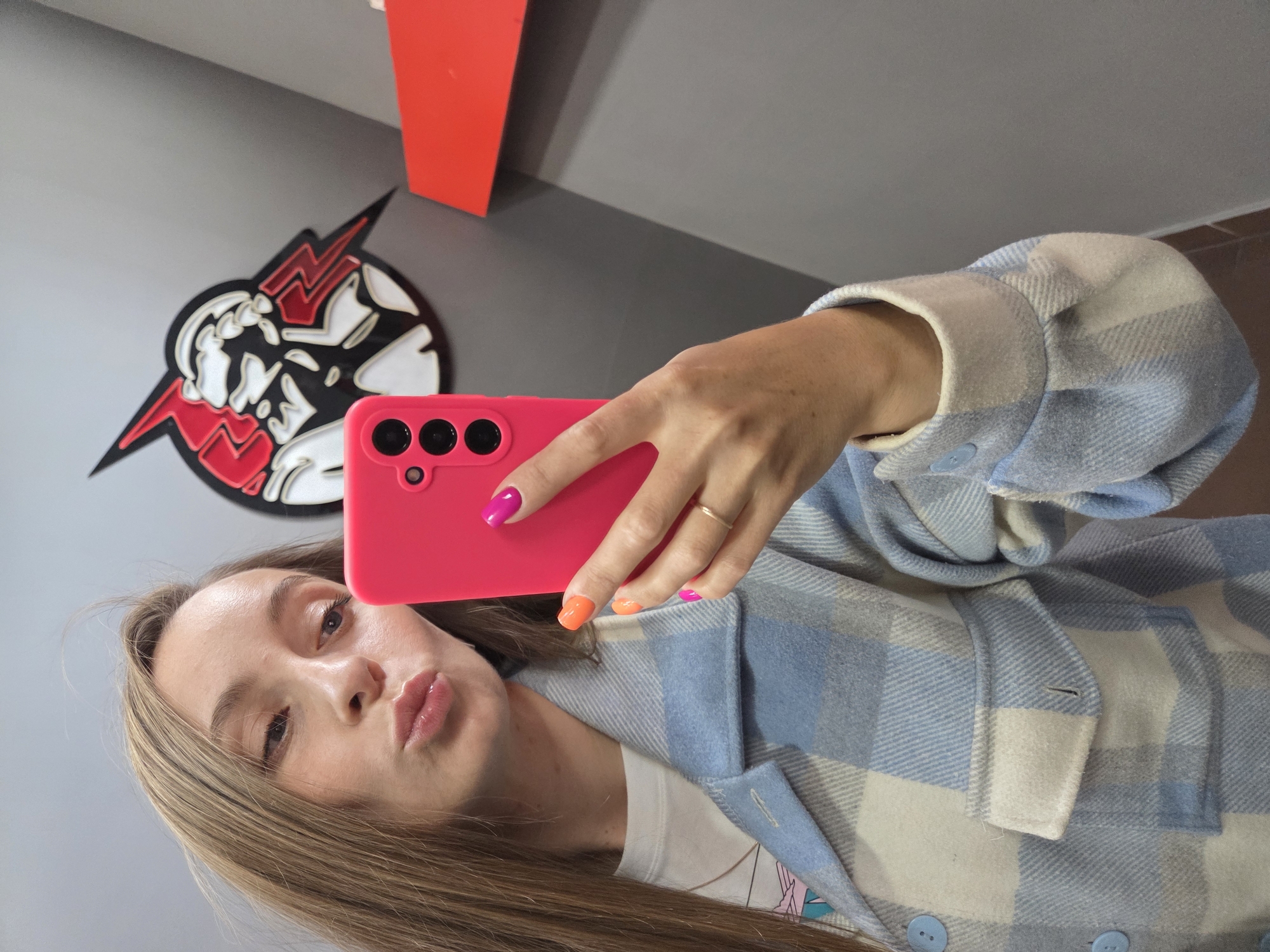 A woman is taking a selfie with a pink smartphone. She has long brown hair and is wearing a blue and white plaid jacket. Her nails are painted pink and orange. In the background, there is a gray wall with a red geometric shape and a black and white logo with red lightning bolts. The location is unknown and there is no verifiable connection to Birobidzhan, Russia.