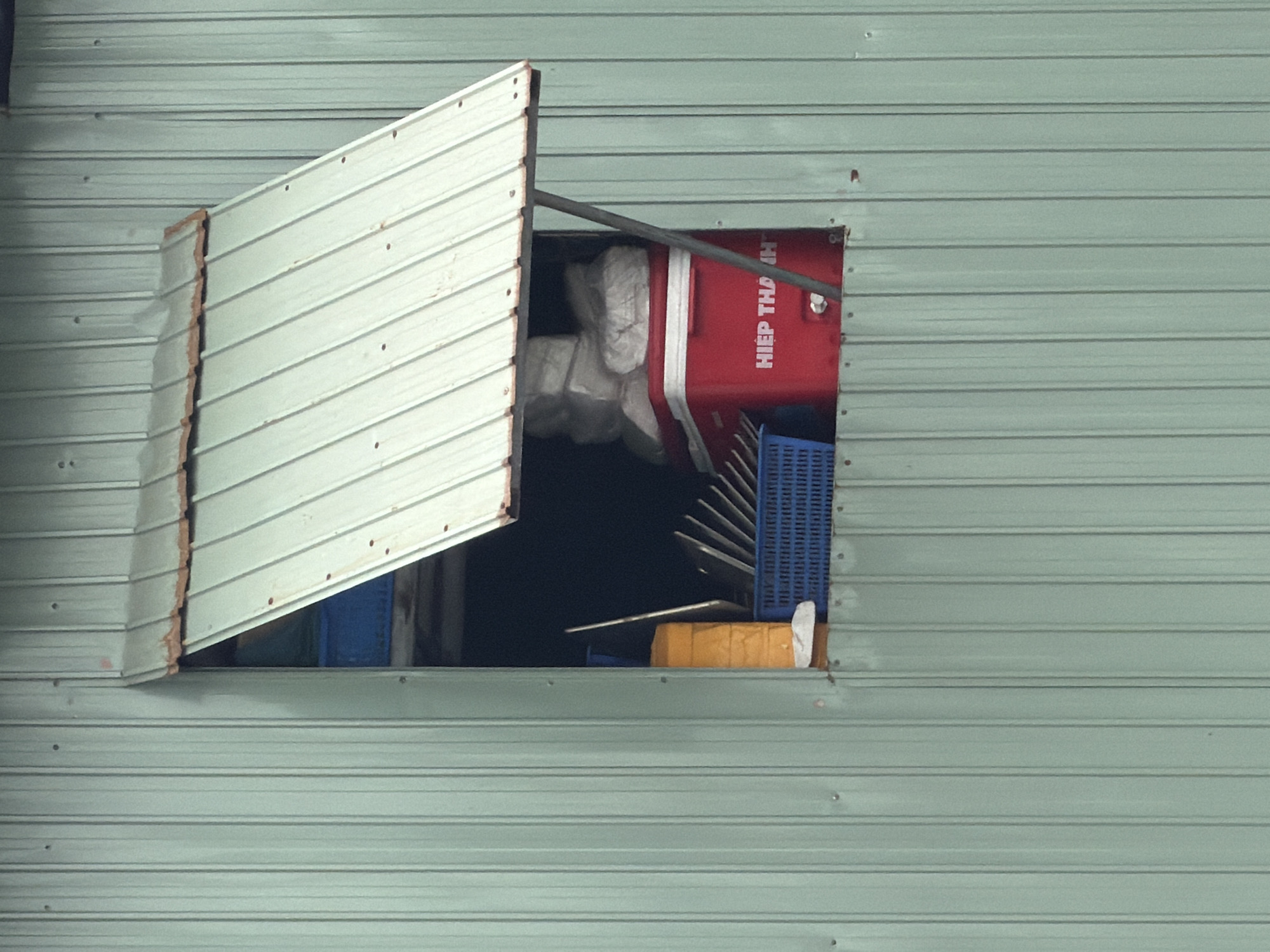 An exterior shot depicts a shed or building in Đà Nẵng, Vietnam, with a partially open window. The window is a rectangular opening in corrugated metal siding painted a light blue-green color. The window has a hinged metal flap, also corrugated and painted white, which is open and propped up by a metal rod. Visible through the opening are various objects, including a red cooler with white straps and the inscription "HIEP THA NH," blue plastic crates, and some white packaging materials. The interior of the shed appears dark. The overall setting is simple and utilitarian, with a focus on storage and functionality. There is no indication of recent human presence in the shot.