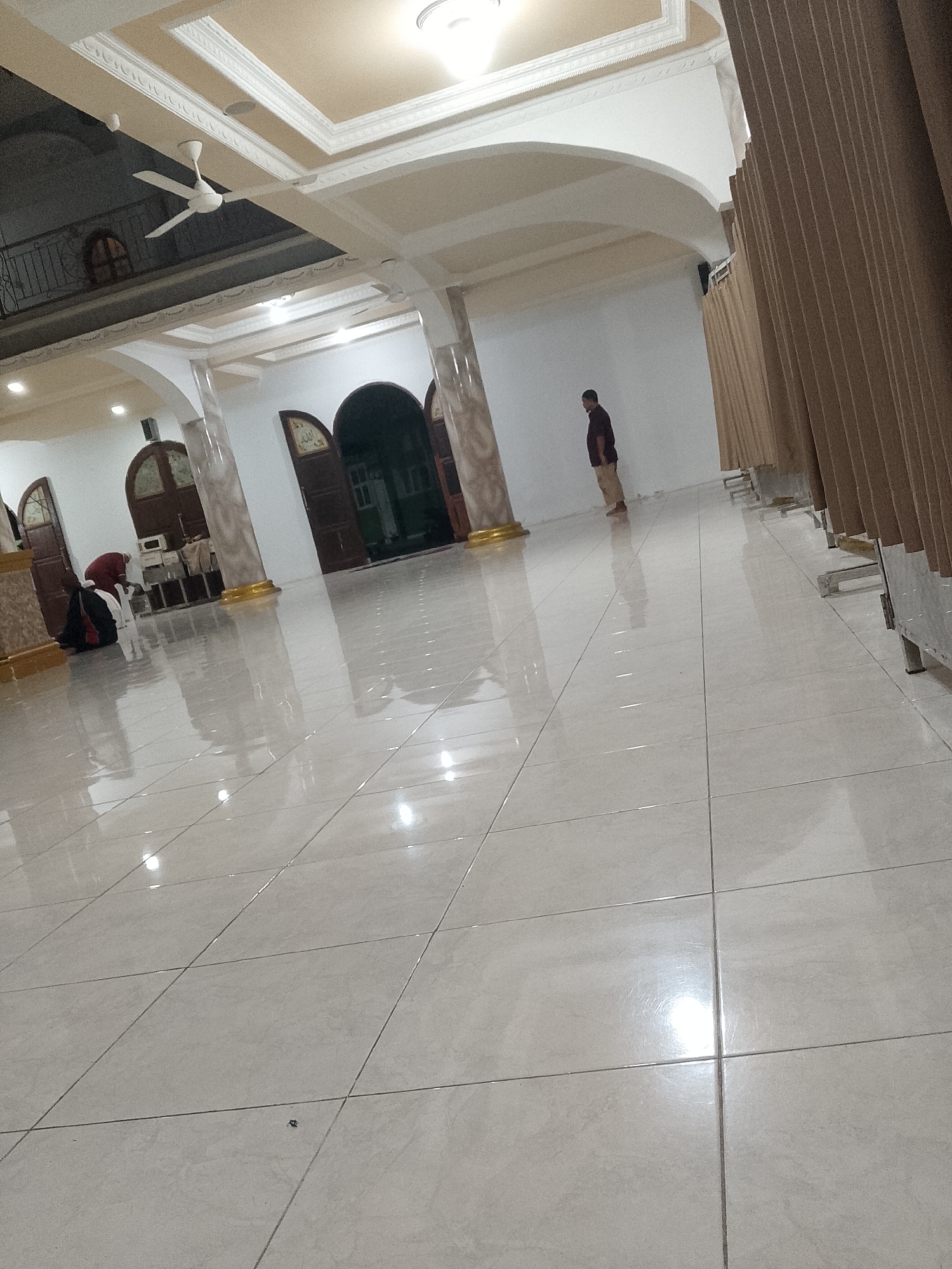 The image depicts the interior of a mosque in Ranoyapo, Indonesia. The floor is made of large, light-colored tiles that reflect the overhead lighting, creating a polished and spacious appearance. A ceiling fan is visible near the ceiling, and decorative molding lines the ceiling's edges. Several large, arched doorways lead into different sections of the mosque. In the background, two individuals are present. One person, wearing a dark shirt and light-colored pants, stands near a draped partition. Another person, wearing a red hijab and dark clothing, is seated on the floor near one of the doorways. The overall scene suggests a quiet and serene environment within the mosque.