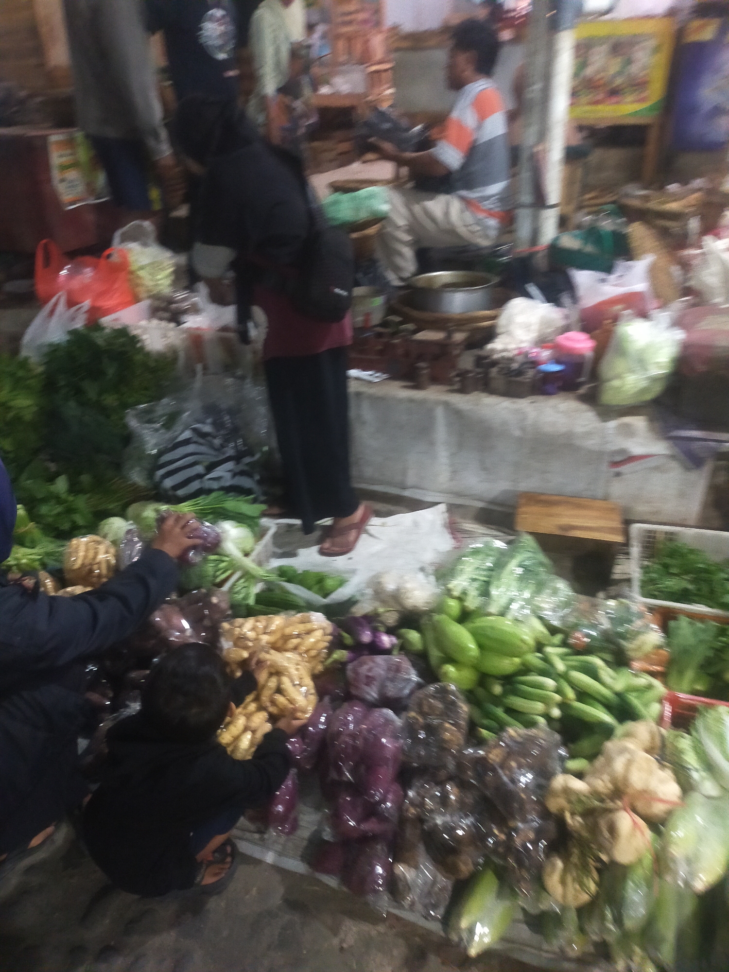 This is a medium-shot, outdoor photograph taken at a market in Trayeman, Indonesia, likely at dusk or in low light conditions given the visible blur and grain. In the foreground, a child wearing a black hoodie and dark pants is crouching, reaching towards a pile of ginger root that is bundled and wrapped in plastic. To the left of the child, a person's arm, also wearing a dark sleeve, is visible, seemingly interacting with vegetables. A variety of produce is displayed on a table in front of them, including what appears to be ginger, eggplant, cucumbers, and leafy greens, some of which are packaged in plastic. In the midground, a person in a black hijab and maroon dress is standing, and another individual in a grey and orange striped shirt and white pants is seated. The background reveals market stalls with various goods, some blurry shelves, and a poster on the wall. The overall atmosphere is that of a bustling local market.