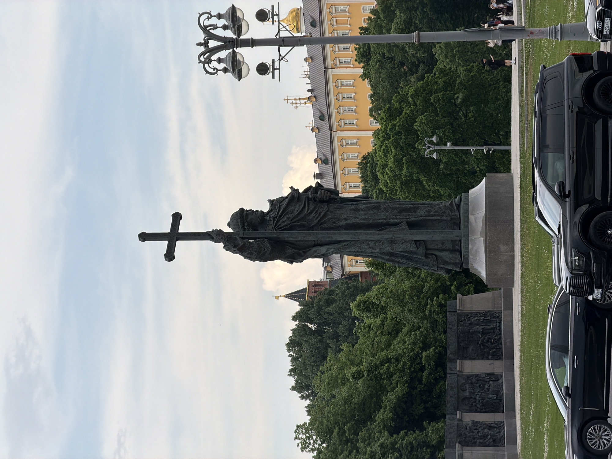 The image shows a statue of a religious figure holding a cross, likely located in Moscow, Russia. The statue stands on a carved pedestal, surrounded by trees and green space. In the background, there is a yellow building with a gold dome, as well as a reddish-brown tower with a golden spire, hinting at the Kremlin area. A decorative lamppost stands nearby. A black car is parked on the right side of the frame, and people can be seen in the distance on a paved walkway. The sky is pale blue with scattered clouds.