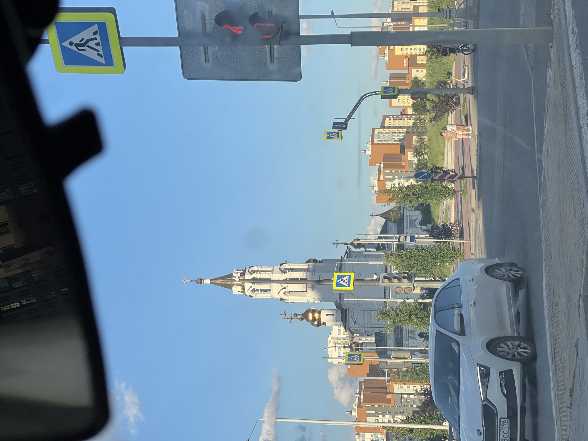 The image captures a street scene in Yekaterinburg, Russia, under a bright blue sky with scattered clouds. In the foreground, a white Skoda sedan is parked. To the right of the car, a traffic light displays a red signal. Further down the street, a white church with golden domes stands prominently, flanked by modern residential buildings in shades of orange and yellow. Several pedestrian crossing signs are visible, one featuring a blue square with a white walking figure inside a white triangle, and another with a blue circle and a white diagonal bar. Pedestrians are seen in the midground, near the church. The environment appears to be a developed urban area, likely an intersection, with trees and greenery interspersed with the buildings.