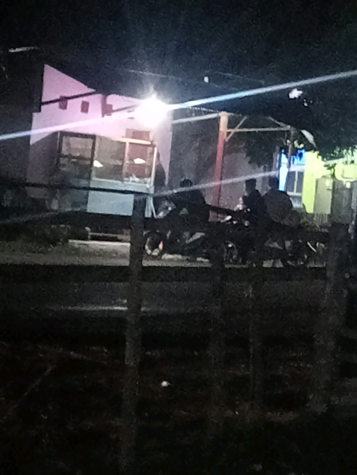 The image depicts an outdoor scene at night in Tanjung Pandan, Indonesia, likely on a street or near a residential area. A building with a white, slanted roof and exposed beams is visible on the left, illuminated by a bright light source, possibly a street lamp, causing a glare across the image. A section of the building has windows or openings that are lit from within. In front of the building, several motorcycles are parked. A group of individuals, silhouetted and indistinct due to low light, are gathered around the motorcycles and in the vicinity of the building. Some are standing, while others appear to be seated. The ground appears to be unpaved and rough, with a fence made of vertical wooden posts in the foreground. The overall atmosphere is dimly lit and suggests a casual gathering or stopover.