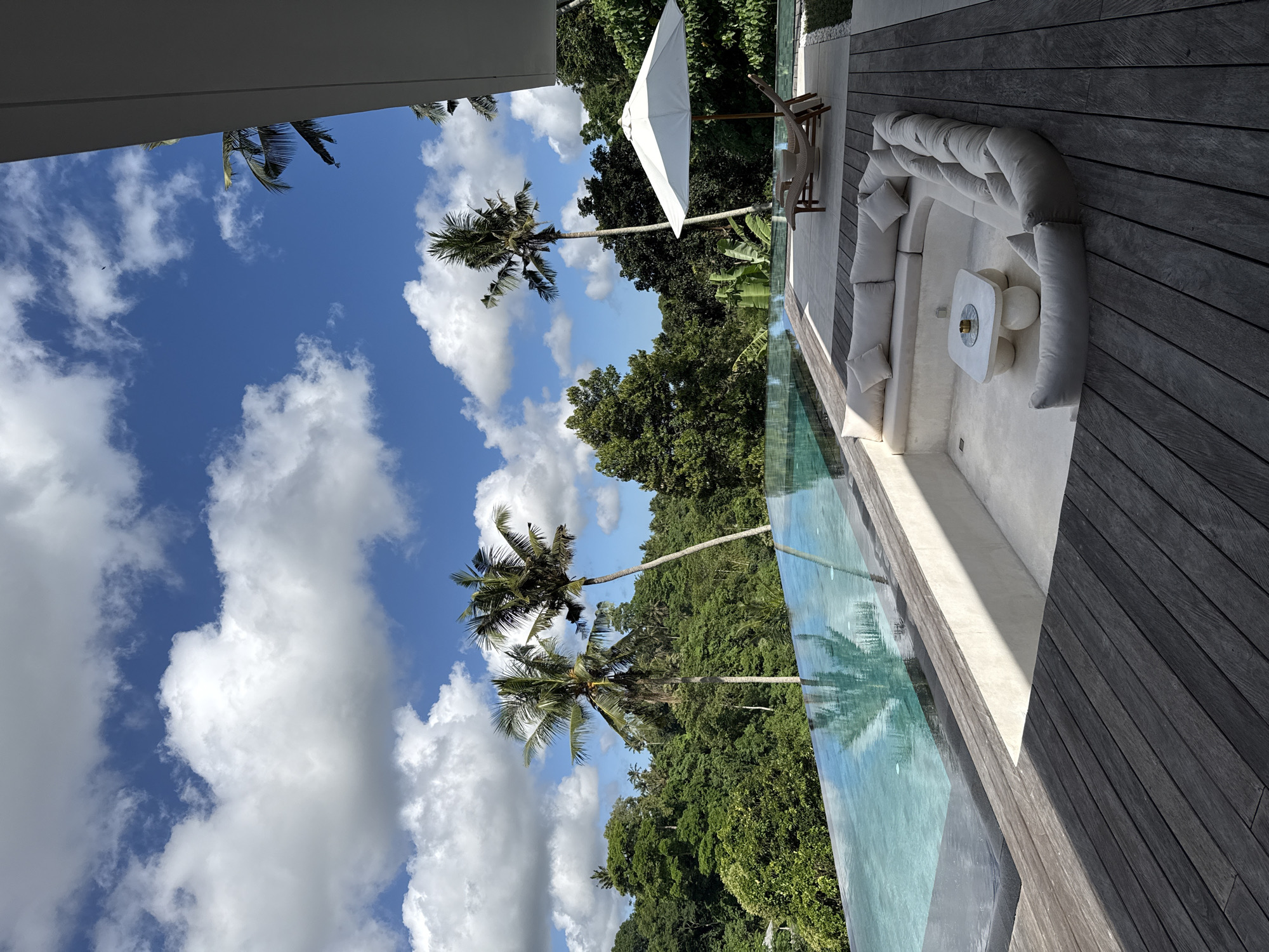 The image shows a luxurious outdoor setting in the Ubud District of Indonesia. The view is of a swimming pool area, adjacent to a grey wooden deck with a large, cream-colored, curved outdoor sofa and a small, white table. There is also a white parasol shading two chairs. Beyond the pool, there is lush green vegetation. The sky is blue with scattered white clouds, and several palm trees are visible. The scene appears to be taken from inside a structure, looking outwards. The pool water is clear and reflects the trees and sky. There is no indication of people or activity in the image.