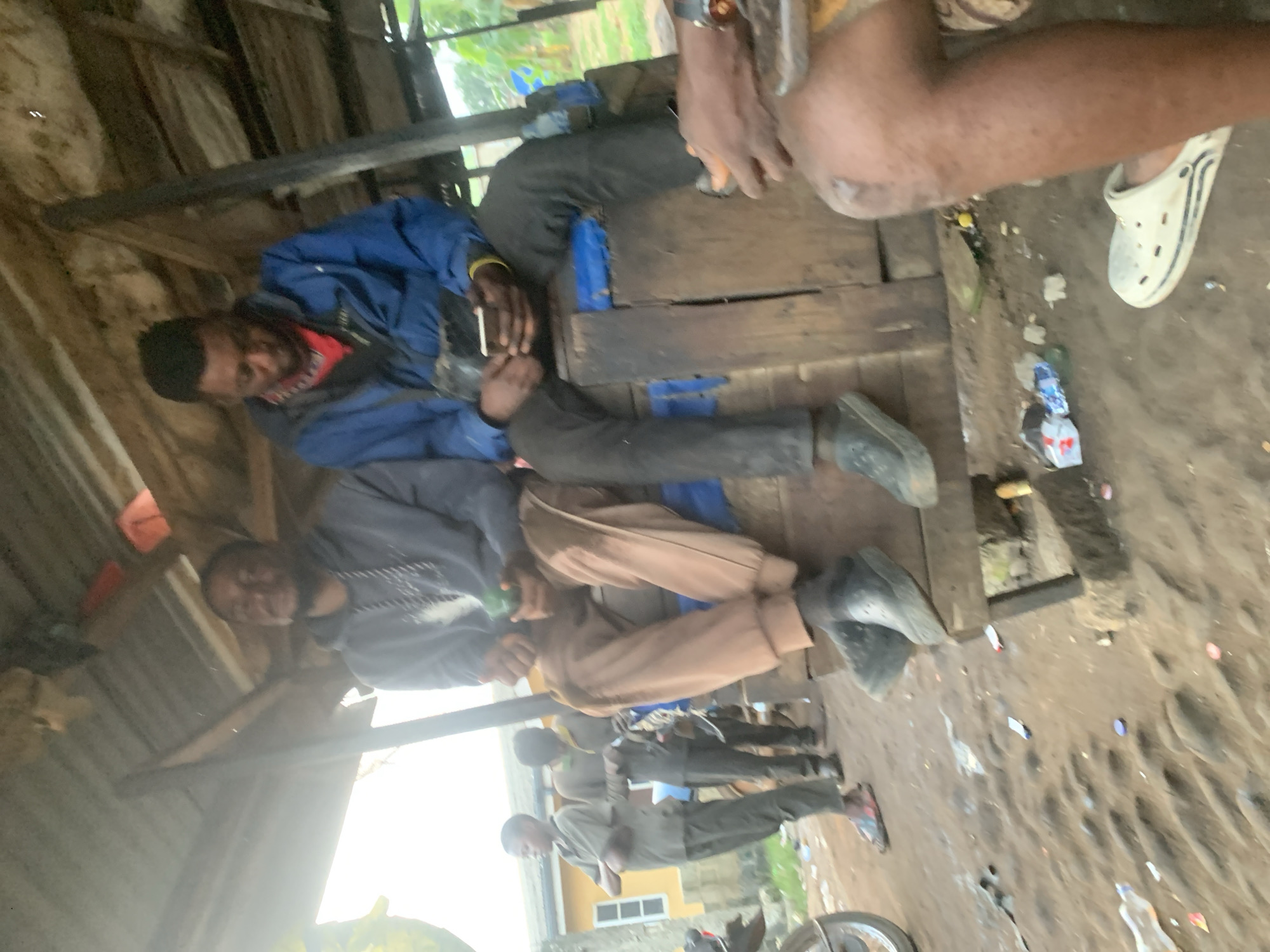 The image depicts an outdoor scene in what appears to be a bustling, informal marketplace or gathering area in Shagamu, Nigeria. Two young men are seated on a wooden structure, likely a bench or makeshift seating. The man on the left is wearing a grey hooded sweatshirt and light brown pants, holding a green object in his right hand. The man on the right is wearing a blue jacket over a red shirt and black pants, actively using a mobile phone with both hands. Their posture suggests a relaxed interaction or shared moment.
In the background, other individuals can be seen, including a young boy in a light brown shirt standing with his arms crossed, and another person further back in a darker garment. The environment is characterized by a rustic, open-air shelter with a corrugated metal roof supported by wooden beams. The ground is uneven and appears to be dirt, scattered with debris including plastic bottles, wrappers, and bottle caps, indicative of a well-used public space. The overall atmosphere suggests a casual, everyday scene in a developing urban setting, with evidence of informal commerce or social gathering.