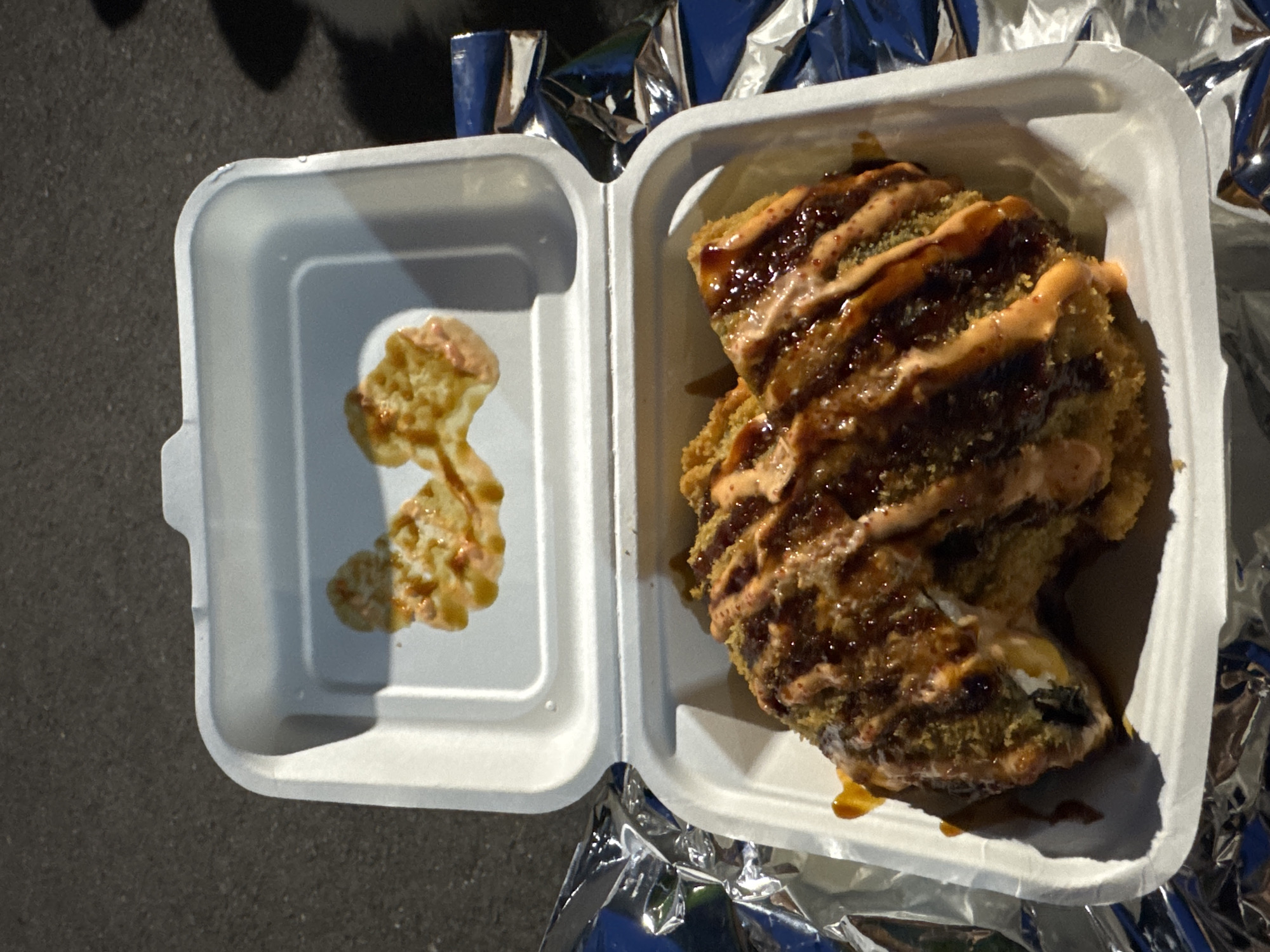 A close-up, overhead shot shows a takeout container filled with a large, deep-fried sushi roll. The roll is coated in breadcrumbs and drizzled with a dark sauce and a creamy, orange sauce. The lid of the container, which is open and placed behind the roll, has two splatters of the orange sauce. The container is placed on a surface covered with crumpled aluminum foil. The background is a dark, asphalt-like surface. The image appears to be taken outdoors, possibly on a street or pavement, with the sun casting a shadow to the right. There are no people visible in the image. The scene is set in Burgas, Bulgaria.