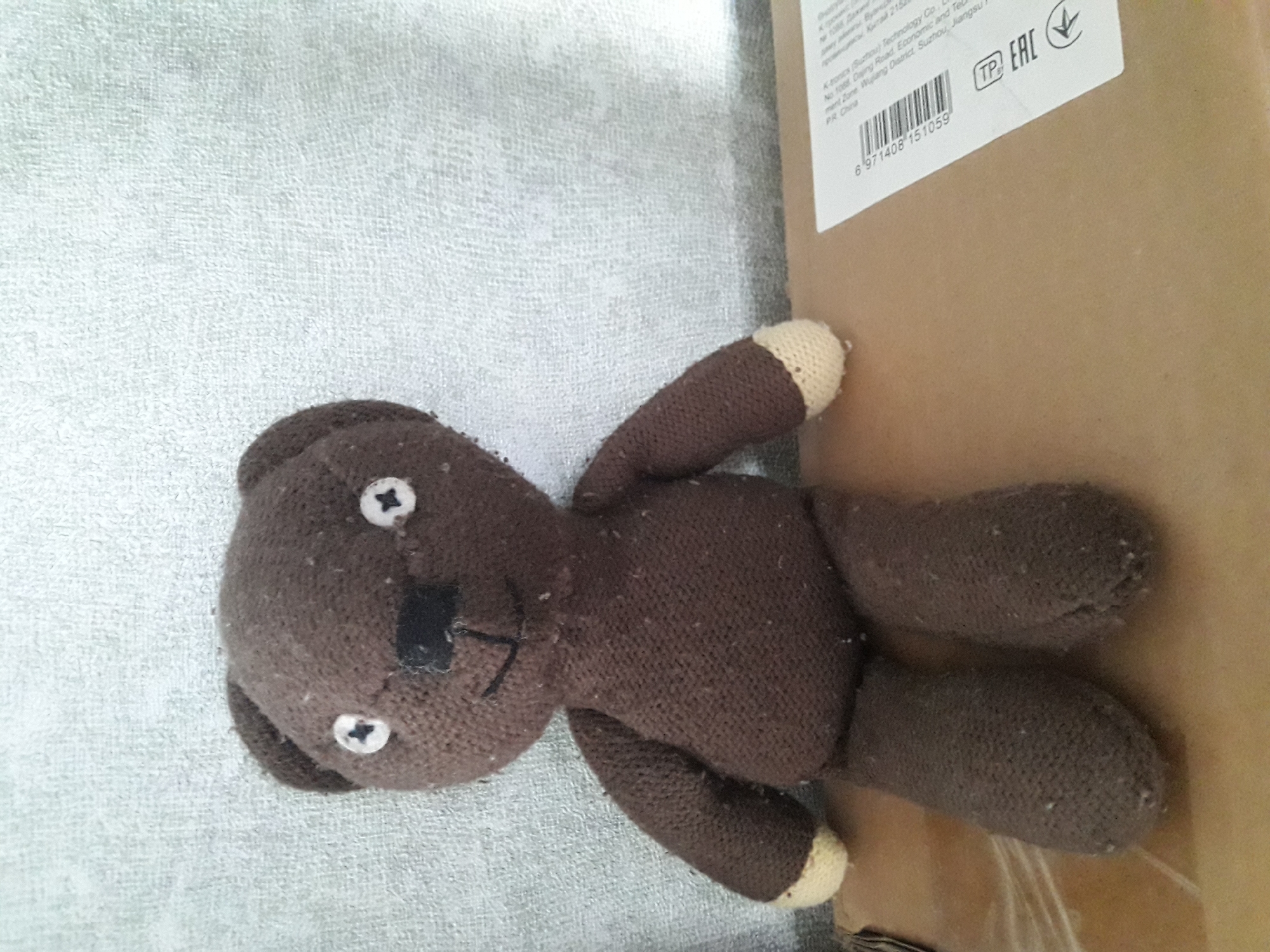 A brown knitted teddy bear with white button eyes and a black stitched nose and mouth is sitting upright on a cardboard surface. The bear is facing forward and appears to be a toy. The background is a textured, light-colored wall. A label with a barcode and some Chinese text is visible on the right side of the cardboard. The scene is indoors, and the lighting suggests it is daytime.