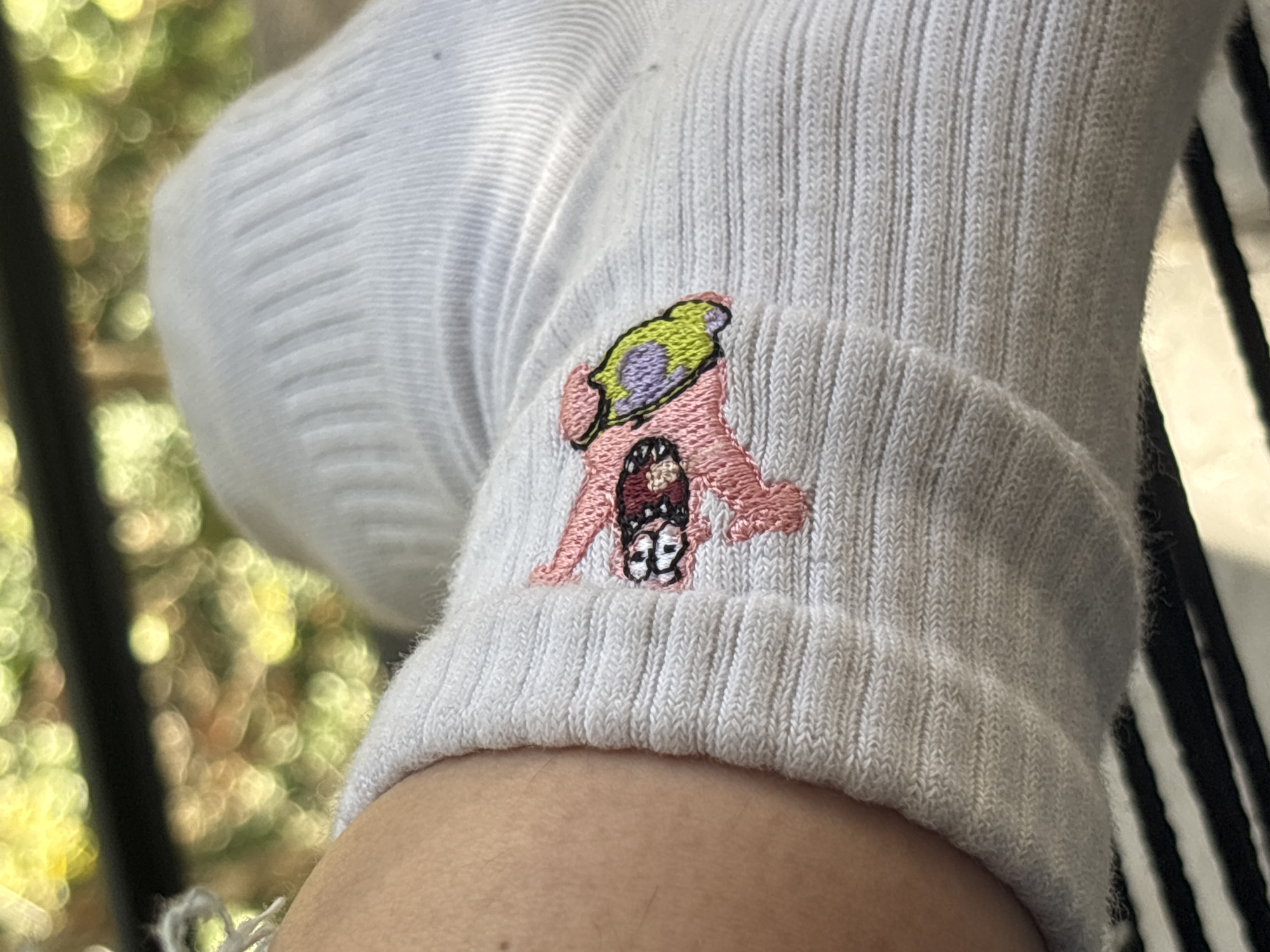 A close-up shot focuses on a person's lower leg, ankle, and foot, prominently featuring a white ribbed sock. The sock is made of a textured, woven material with vertical stripes. An embroidered cartoon character adorns the side of the sock. This character is predominantly pink, with a distinctive light green and purple element on its head or top, resembling a hat or an abstract shape. Its face features black and white details, possibly depicting eyes and a mouth.
The background is softly blurred with a bokeh effect, showing bright, diffused patches of green and yellow, suggesting foliage or trees, indicating an outdoor setting or a bright indoor area with a view of greenery. A dark vertical element is visible on the left side of the background, and a striped object, possibly part of a shoe or furniture, can be seen on the right. The lighting is bright, suggesting it is daytime. There is no visible text or specific activity taking place, rather a static detail shot. The setting is implied to be in Buenos Aires, Argentina, though no specific visual cues confirm this location.