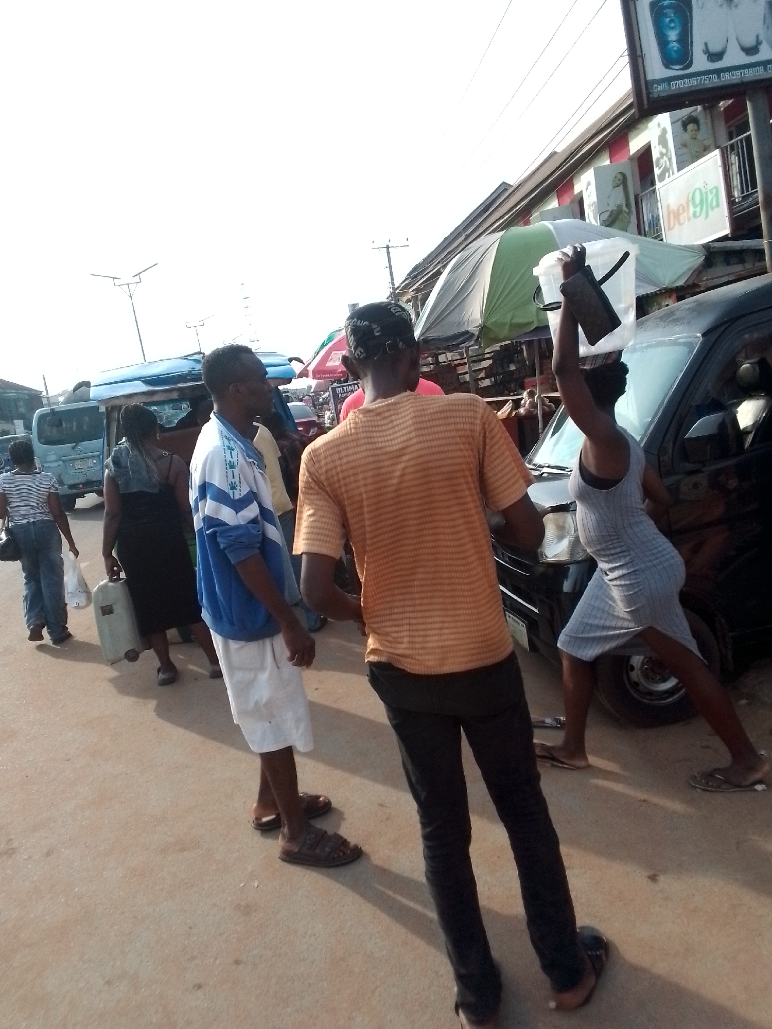 The image depicts a street scene in Orlu, Nigeria. Several people are present, including a woman in a gray dress holding a black object over a black minivan, two men in casual clothing standing nearby, and other individuals walking along the street. The environment includes buildings, utility poles, and vehicles, suggesting a commercial or residential area. A “bet9ja” sign is visible on a building. The setting appears to be a bustling street or marketplace.
