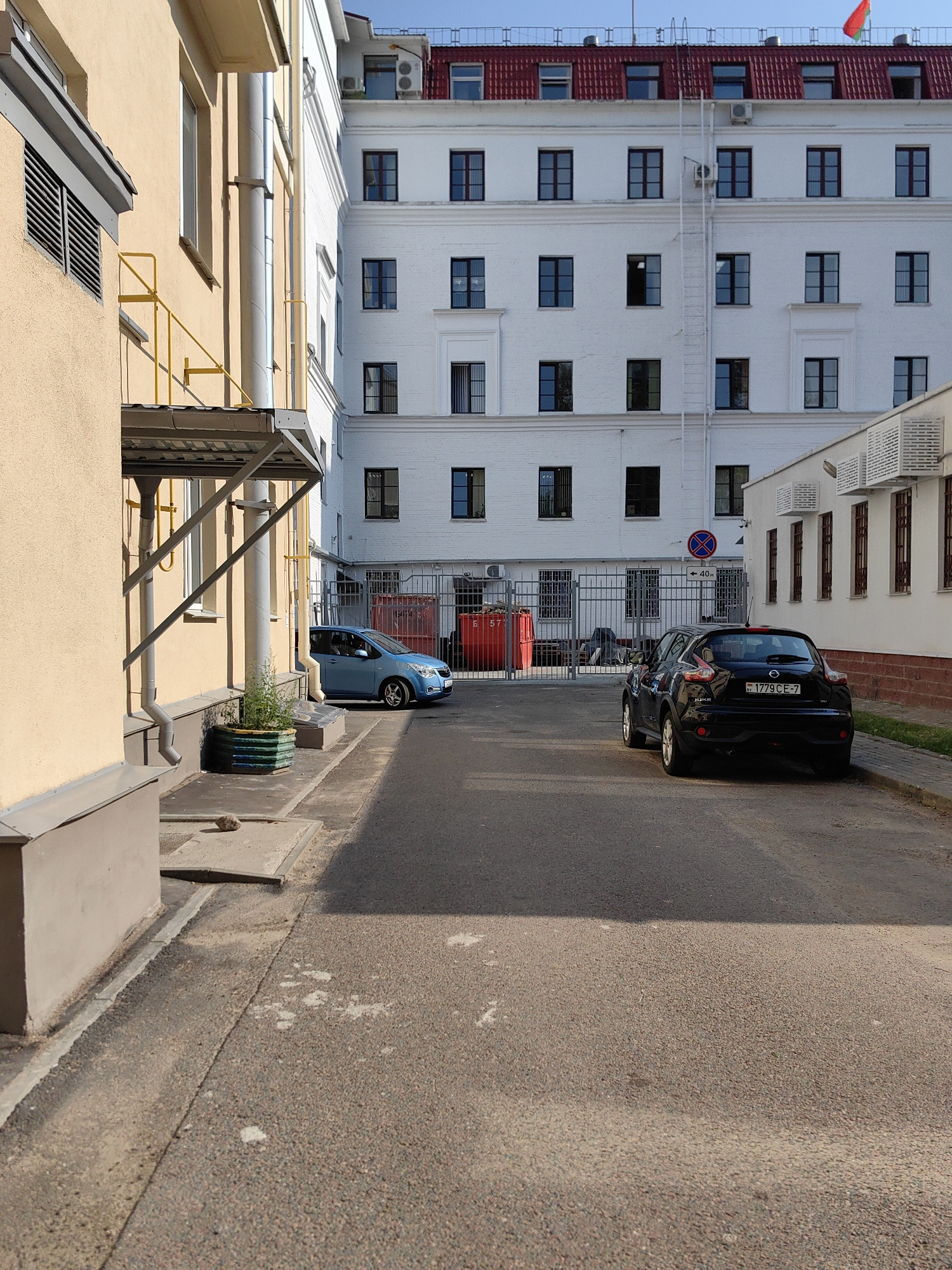 A black Nissan Juke is parked on the right side of an asphalt road, facing away from the camera. The car's license plate reads "BY 1779 CE-7". To the left of the Nissan, a blue compact car is parked on a sidewalk. Behind the cars, a white building with multiple windows is visible. The building has a red roof and a flag pole with a red and green flag at the top. Further to the left, a yellow metal railing extends from a beige building. The scene appears to be an urban environment, possibly a courtyard or alleyway, in Minsk, Belarus.