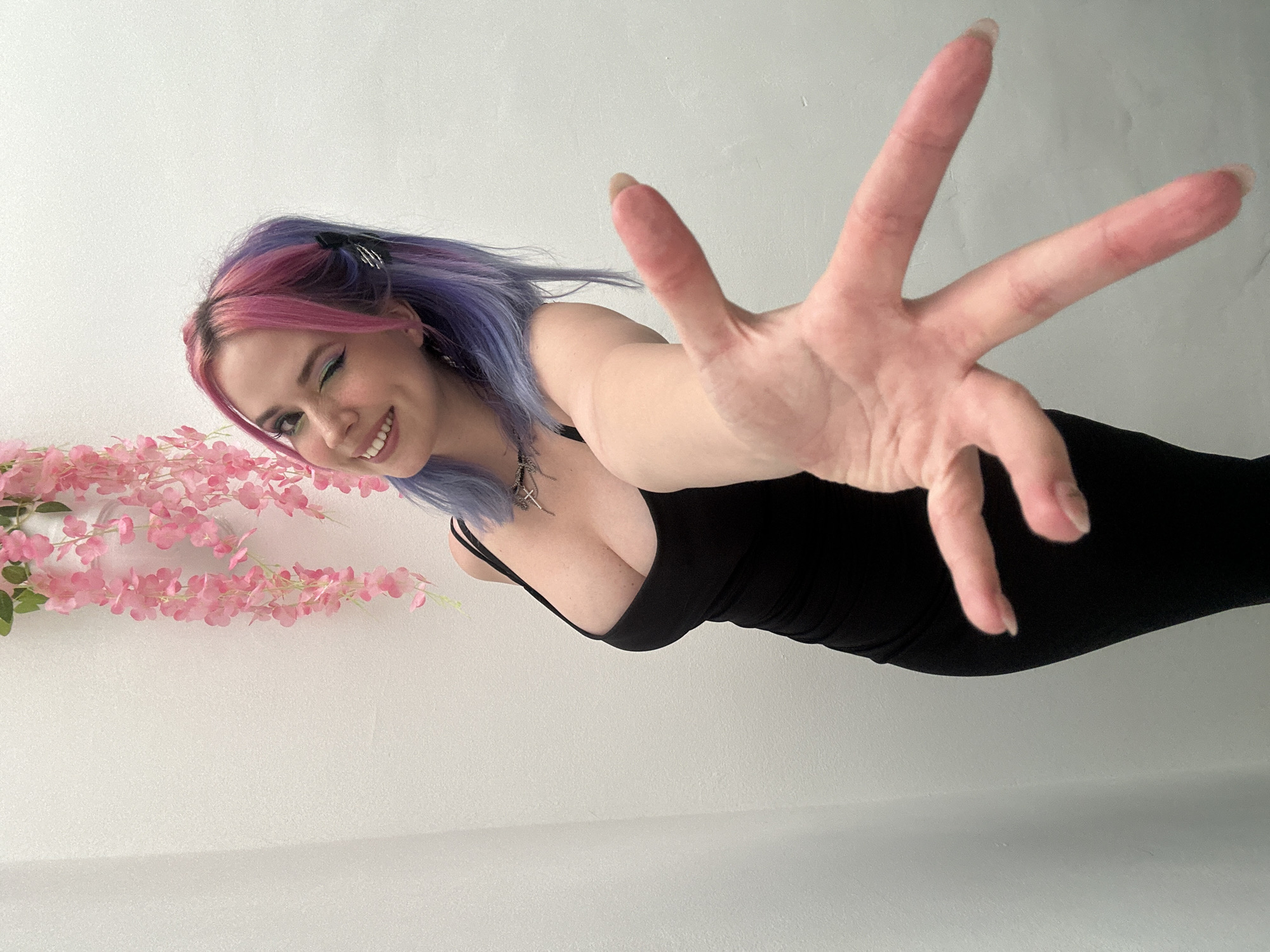 A young woman is pictured indoors against a white wall, with a strand of pink flowers to the left of the frame. She has pink and blue hair, a black dress, and is reaching towards the camera with her right hand fully extended. She is smiling. It is not possible to verify the location in City of Zagreb, Croatia from the image.