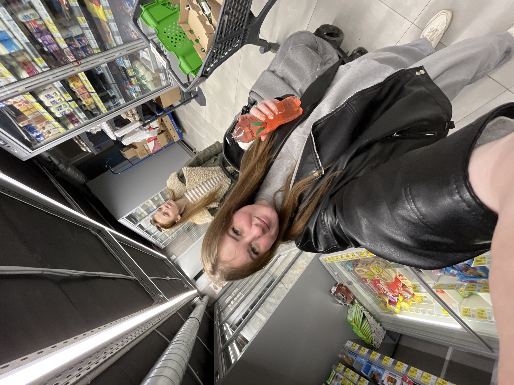 A selfie taken in a supermarket aisle. A young woman in a black leather jacket and grey sweatpants holds a bottle of orange liquid. Beside her is a shopping cart with a green basket. A second woman is visible further down the aisle, standing near refrigerated shelves. The shelves contain various food items. The setting is a grocery store with fluorescent lighting. The location is presumed to be Chaikovsky, Russia, based on additional context.