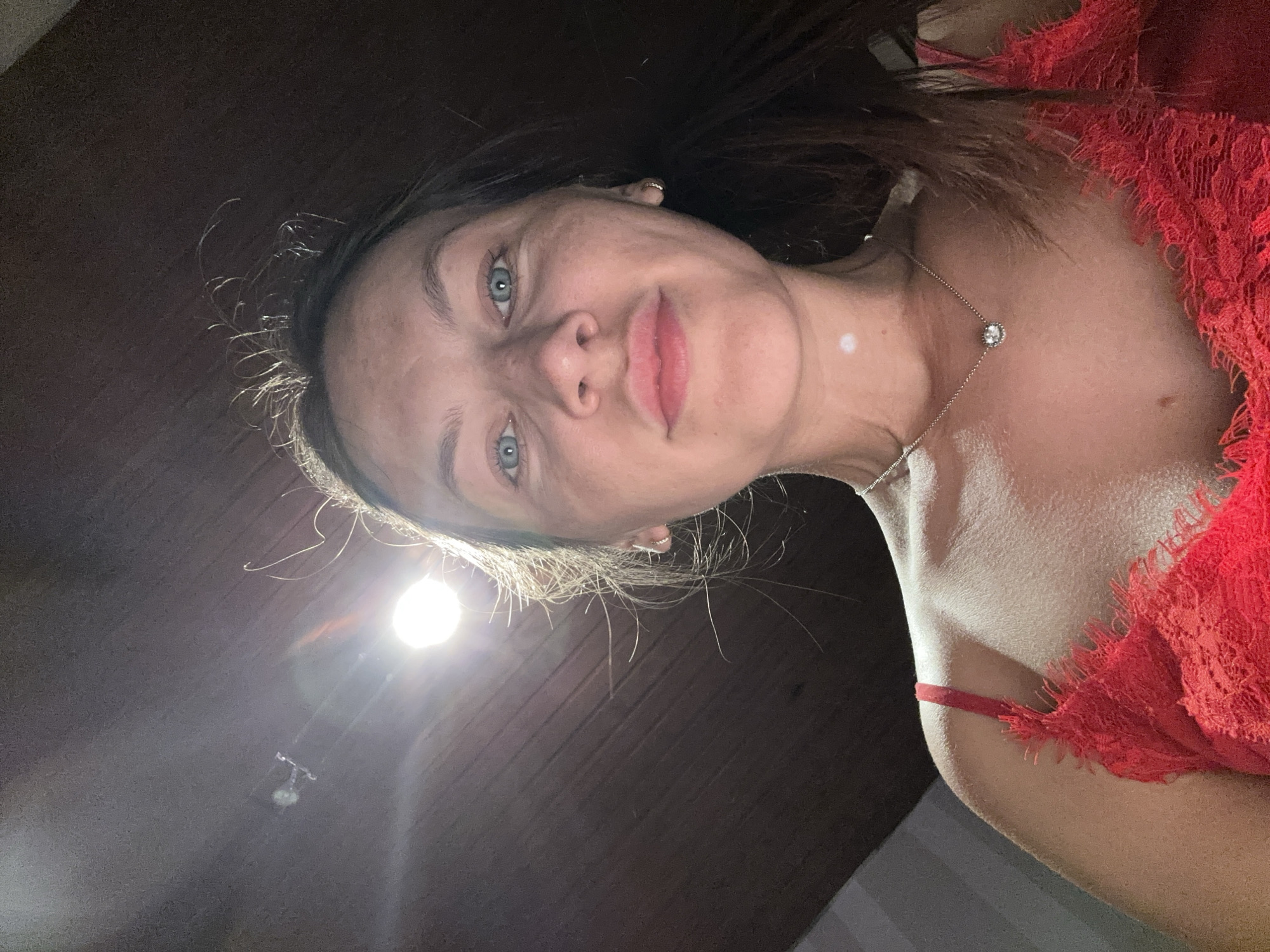 A woman with fair skin, blue eyes, and dark hair is depicted from a low-angle perspective. She wears a red, lace-trimmed garment and a delicate necklace with a single stone. The background is dark, but a light fixture is visible above her. There's no information available about this image being in Fredenbeck, Germany.