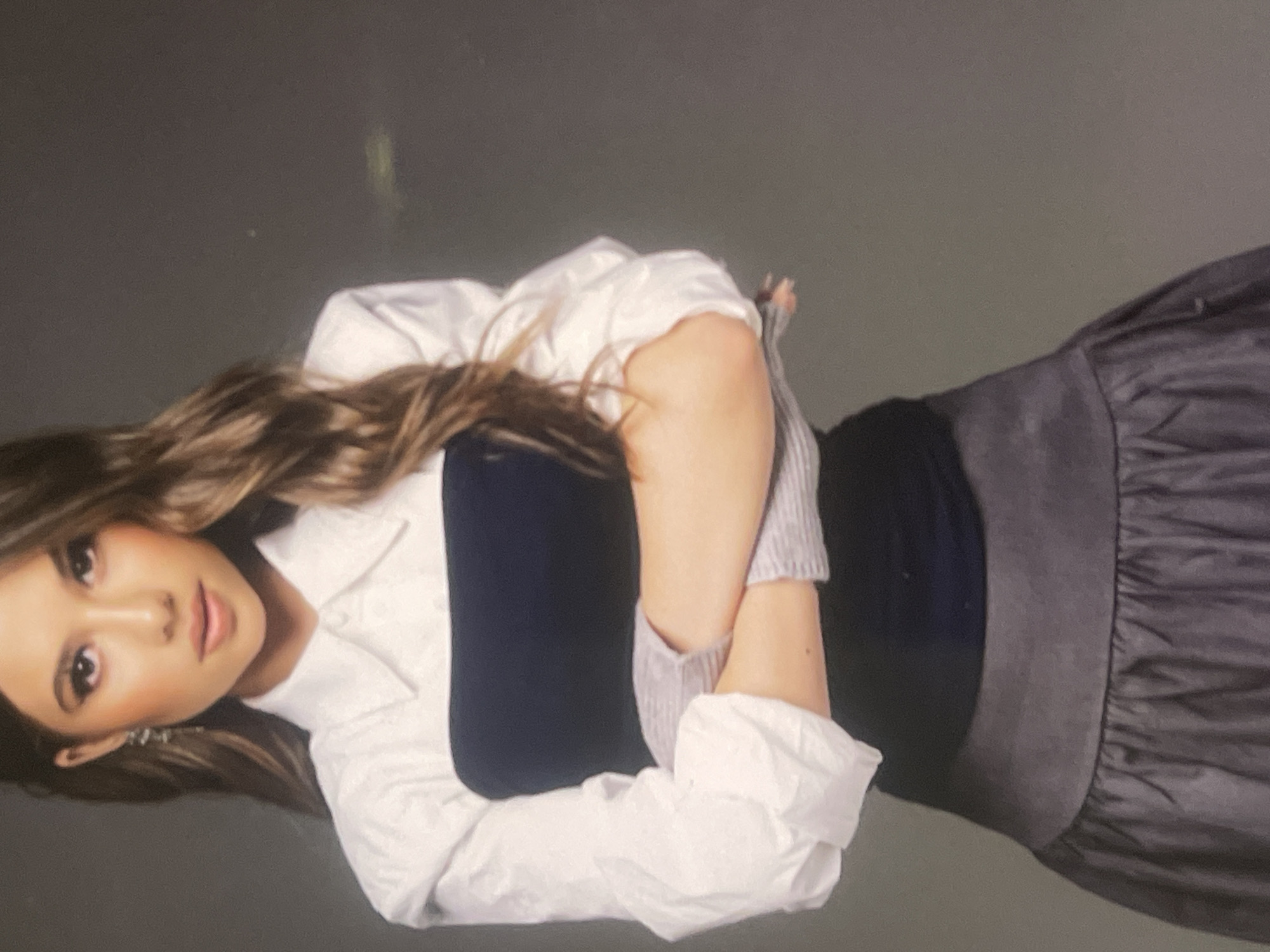 A portrait photograph captures a young woman posing against a plain, dark grey backdrop, likely indoors. She is positioned facing forward, looking directly at the viewer with a neutral and composed expression. Her long, wavy, dark brown hair is parted on the side and falls over her shoulders.
She is dressed in a white collared shirt with the sleeves pushed up to her forearms, revealing what appears to be light grey, ribbed cuffs, possibly from a sweater worn underneath or fingerless gloves. Beneath the white shirt, she wears a dark navy or black sleeveless top. Her arms are crossed over her chest. Completing her outfit is a dark grey or black skirt, featuring gathered or ruched detailing on the right side, with a flat waistband.
Her facial features include soft makeup, emphasizing her eyes and light pink lips. A subtle, metallic earring is visible on her left ear. The lighting is soft and even, highlighting her features without harsh shadows. There are no visible location cues within the image to specify Poltava, Ukraine, beyond the provided context. No text is visible in the image. The scene depicts a posed moment, possibly for a fashion or personal photoshoot.