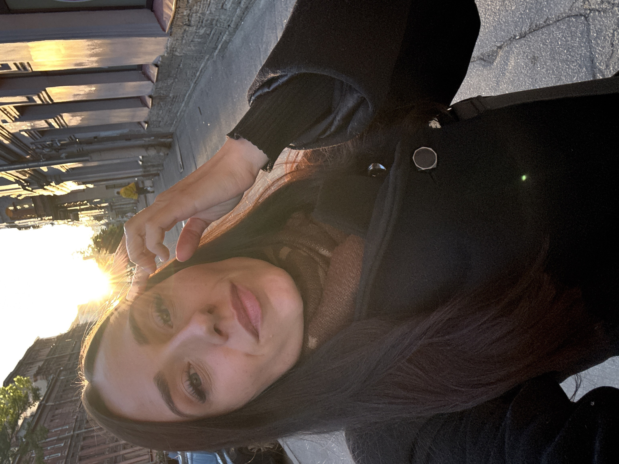 A woman takes a selfie on a sidewalk in Saint Petersburg, Russia, during a sunset. The low sun casts long shadows and creates lens flare across the image. The woman is wearing a dark coat and a patterned scarf. In the background, the facades of old brick buildings line the street, and a person in a yellow jacket walks away from the camera. The pavement is cracked and worn.