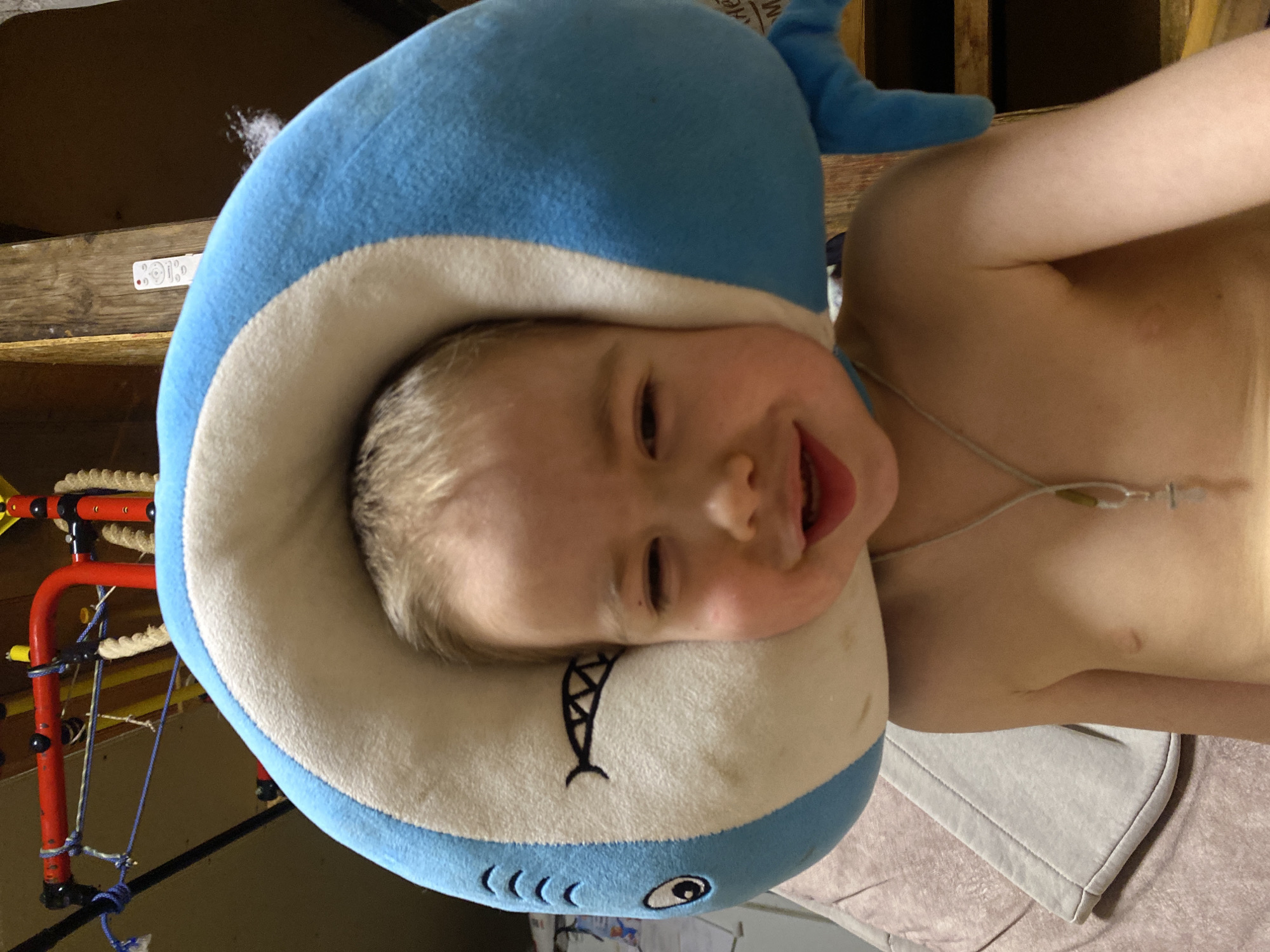 A young child, appearing to be a toddler or preschooler with blonde hair, is lying down and smiling broadly at the camera. The child is wearing a blue and white shark-themed neck pillow or hood, which frames their face. The shark pillow features embroidered eyes, gills, and a mouth. The child's upper torso is visible, suggesting they are shirtless or wearing light-colored clothing. A thin, light-colored medical line or tube with an attachment is visible on the child's chest and neck area.
The background shows an indoor setting with wooden beams and structures, possibly indicating a part of a house or a therapy room. To the left, there is a red and yellow structure, possibly a piece of play or therapy equipment, with ropes and blue cords attached. In the upper background, a white remote control is resting on a wooden surface. A portion of a blanket or towel is visible beneath the child. On a wooden beam in the background, partially visible text reads "NEX". The child appears happy and comfortable.