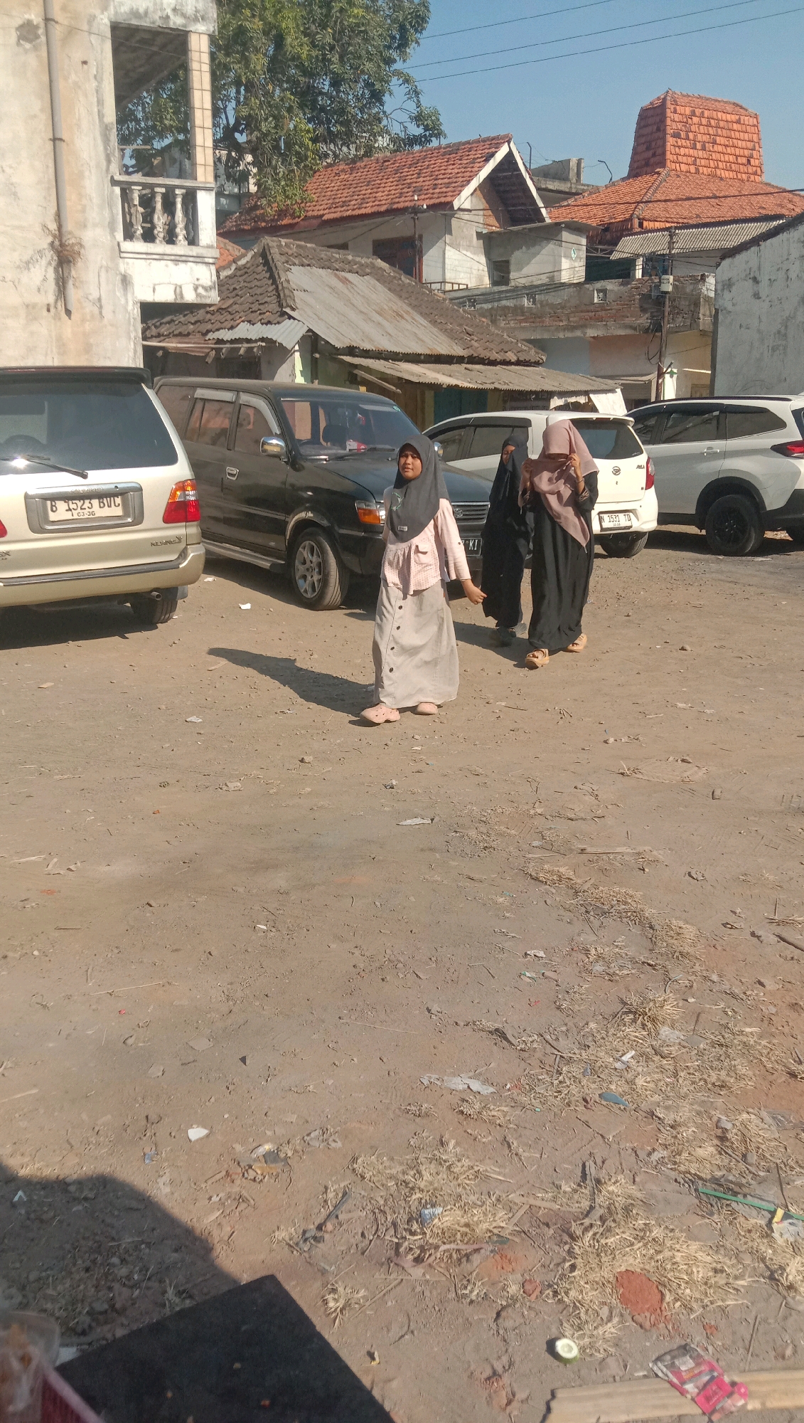 A medium shot shows three women, two wearing hijabs, walking across a dirt lot in Sidogiri, Indonesia. The woman in the foreground is wearing a gray hijab and a long, light gray skirt with a light pink shirt. She is walking from left to right, looking towards the camera. Behind her, two women are walking together, also in hijabs. The woman on the left is wearing a dark hijab and a dark dress, while the woman on the right is wearing a light pink hijab and a dark dress. They are walking in the same direction as the first woman. In the background, several cars are parked, including a silver SUV, a black SUV, and a white SUV. Buildings with red-tiled roofs are visible behind the cars. The ground is covered in dirt, with some dry grass and debris scattered around. The sky is clear and blue.