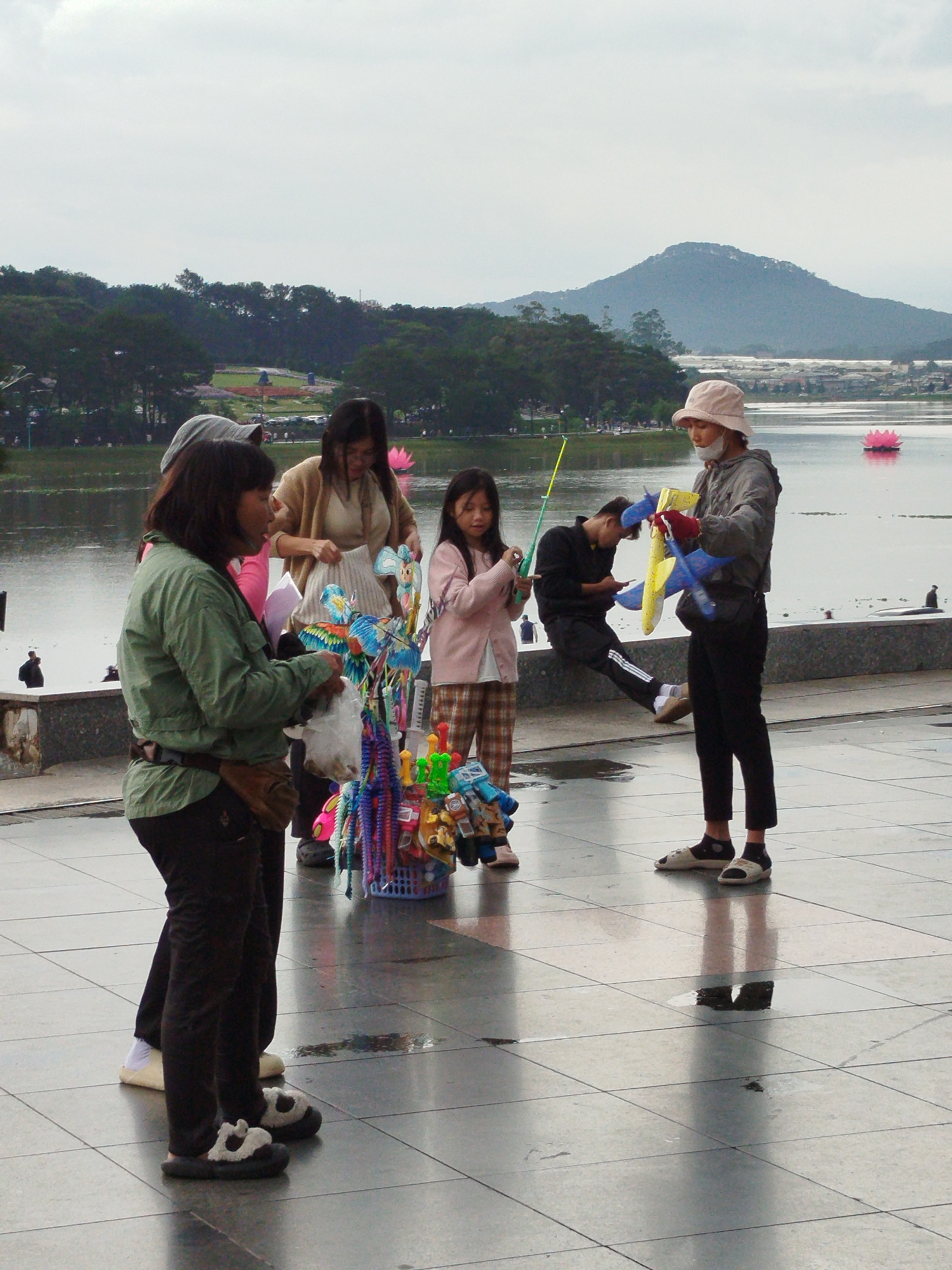 The image shows a vendor in Da Lat, Vietnam, selling toys on a paved plaza near a lake. The vendor, wearing a green jacket and dark pants, stands next to a basket full of colorful toys. She is serving a person wearing a hat and grey jacket. A woman in a tan jacket and a girl in a pink sweater are standing near the toys. A person in black is sitting on the wall looking at a phone. Behind the plaza, there is a lake with a pink lotus flower floating on the surface. Beyond the lake are trees and a mountain in the distance. The sky is overcast, and the plaza surface appears wet, with reflections visible.