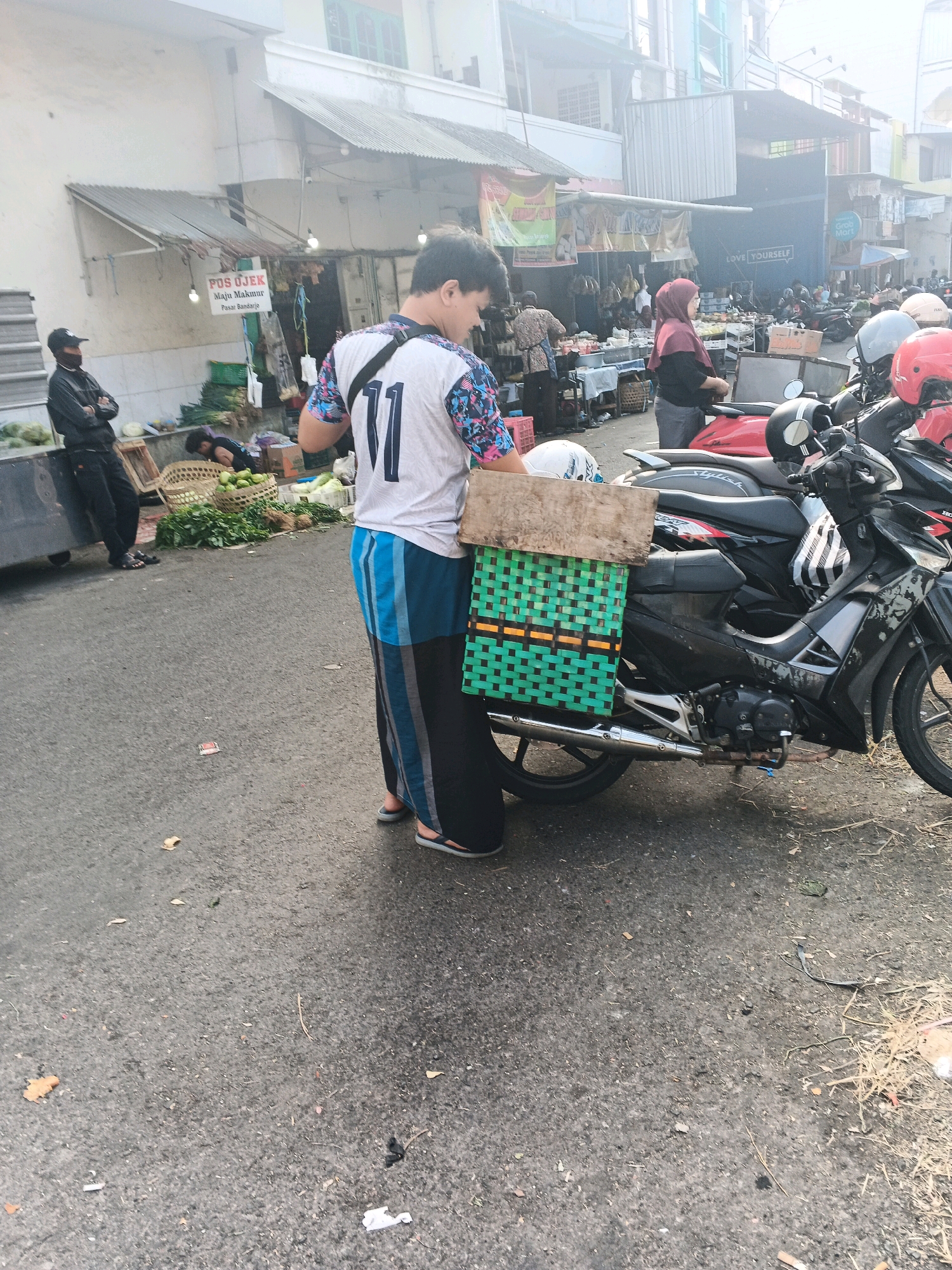 The image depicts a street scene in Ungaran, Indonesia, likely at a market or roadside stall. In the foreground, a young man wearing a white t-shirt with a graphic print and the number "11" on it, along with a sarong, stands beside a parked motorcycle. He has a green woven basket attached to the back of the motorcycle, with a wooden plank on top. The motorcycle is black and gray. In the background, there are several other motorcycles, some with helmets on them, and various stalls with goods, including vegetables, displayed. Several people are visible in the background, some appearing to be vendors and others customers or passersby. A man wearing a black jacket and a face mask is seen standing near a stall to the left. The environment appears to be outdoors, with buildings lining the street and what seems to be a somewhat hazy atmosphere.