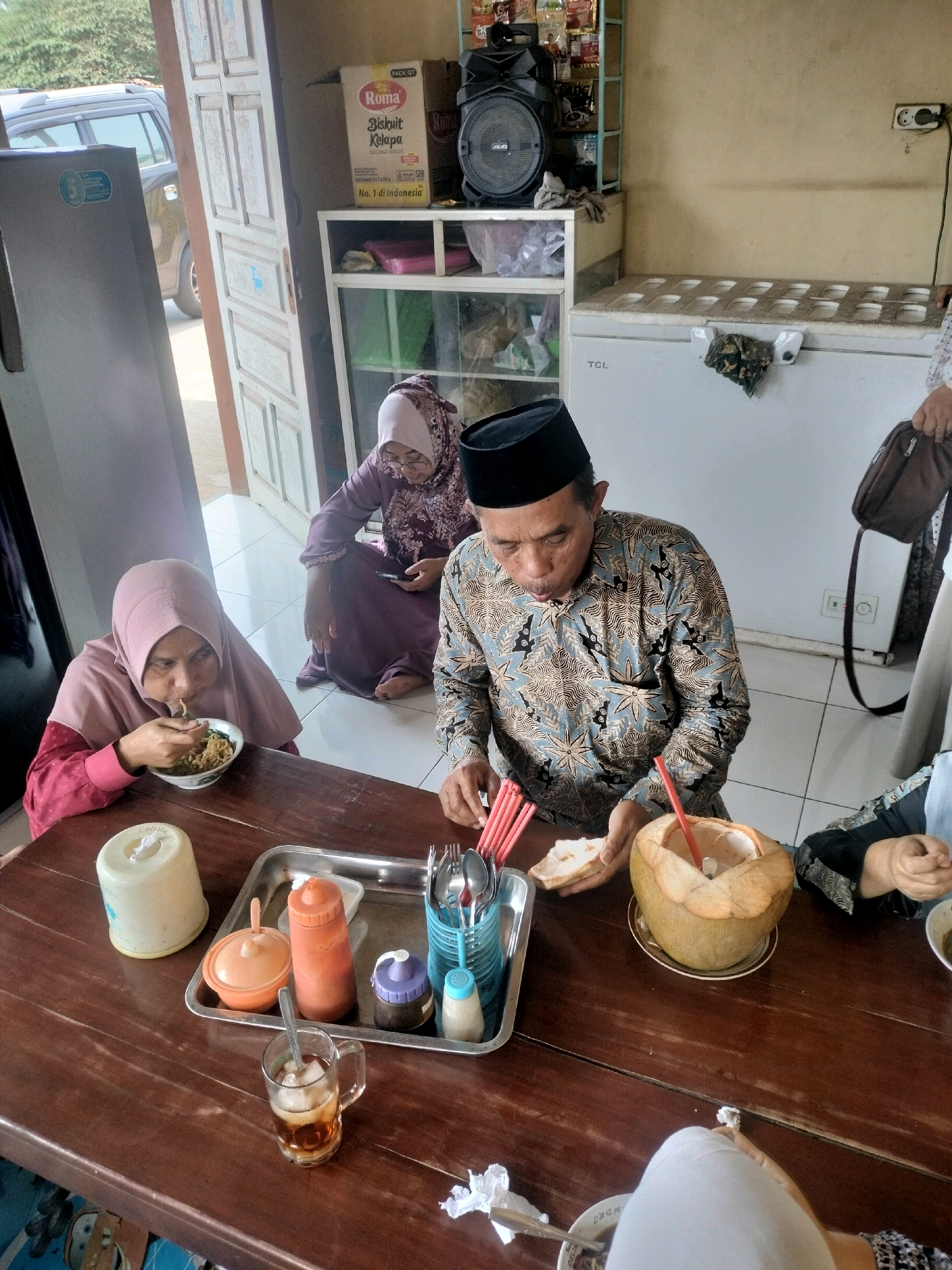 In the city of Kutosari, Indonesia, a group of people are gathered around a wooden table, seemingly in a casual dining setting, possibly a home or a small eatery. The scene is brightly lit by natural light filtering in from the side, suggesting it's daytime.
At the center of the composition, a man wearing a batik shirt and a black peci (skullcap) is actively eating, his mouth open as he consumes food from a bowl. To his left, a woman in a light pink headscarf is also eating noodles from a bowl with a spoon, her gaze focused on her meal. Another woman, further in the background, is seated and wearing a purple dress and a matching headscarf, appearing to be engaged with her phone while also looking towards the table.
The table is laden with various items. In the foreground, a glass of iced tea with a spoon sits prominently. To its right, a whole coconut with a straw inserted serves as a beverage. A metal tray holds a collection of condiments, including a red sauce bottle, a dark sauce bottle, and a small white dispenser, along with cutlery. A tissue box is also present on the table.
The background reveals a rustic interior. A large white refrigerator stands to the right, with a power outlet visible on the wall above it. To the left, a refrigerator and a doorway leading outside are partially visible. In the middle ground, shelves hold various items, including a box of "Roma Biskuit Kelapa" and a large speaker, suggesting a blend of domestic and commercial elements. The flooring appears to be tiled. The overall atmosphere is one of a simple, shared meal.