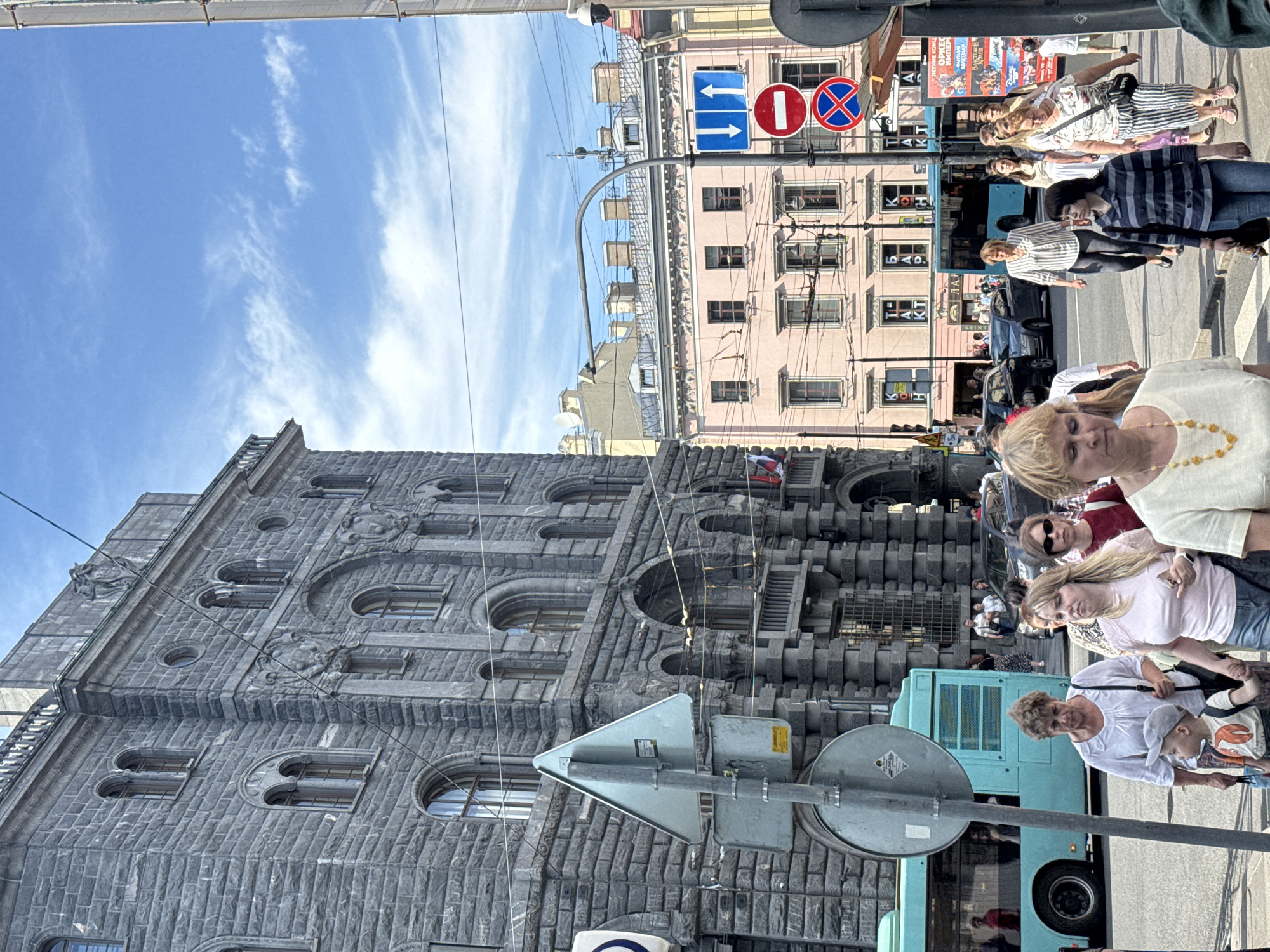 This image depicts a street scene in what appears to be an urban environment, possibly in Europe given the architecture and signage. The perspective is unusual, as the image is oriented vertically, making it seem as though the viewer is looking up at the scene.
The foreground features a large, ornate stone building with distinctive architectural details, including arched windows and decorative carvings. In front of the building, a turquoise bus is visible, along with several people walking and standing. Some individuals are closer to the camera, while others are further away, in the background near the bus. The people are dressed in casual summer attire, suggesting warm weather.
A street sign with directional arrows and prohibitory symbols is prominently positioned in the mid-ground, indicating traffic regulations. In the background, several other buildings with multiple floors and windows can be seen, painted in various pastel colors. The sky above is blue with some wispy white clouds.
The overall impression is a bustling city street during daytime, with people engaged in everyday activities like walking and waiting for transportation. The lighting suggests it is likely midday, with clear skies and bright sunshine, casting shadows. There is no explicit text visible that clearly identifies the location. The emotions of the visible individuals are neutral or difficult to discern due to the distance and angle.