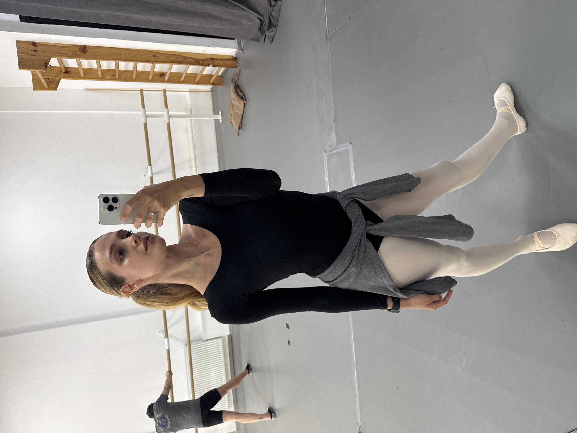 A woman in a black leotard, white tights, and ballet slippers is taking a selfie in a dance studio. She has a gray wrap tied around her waist. Another person, wearing a gray shirt and black shorts, is at a ballet barre in the background. The studio has white walls, a light gray floor, and ballet barres mounted to the walls. There is a wooden stall bar in the background. The studio is located in Tbilisi, Georgia.