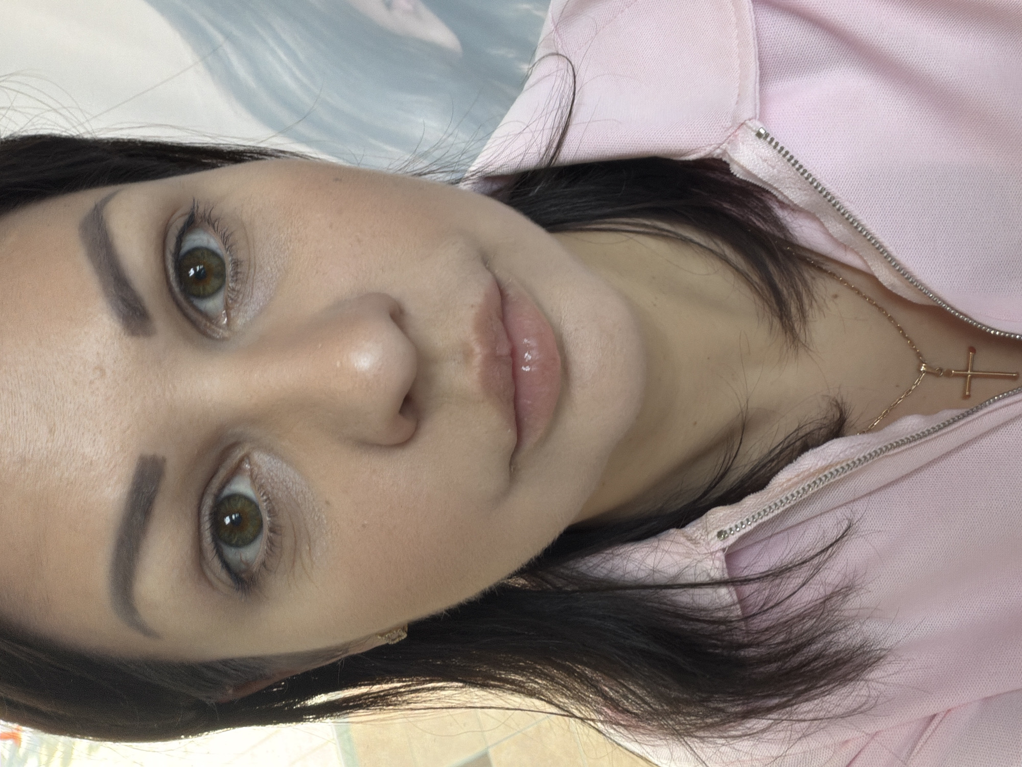 A medium shot captures a woman's face from the chest up, taken outdoors in an urban setting, possibly Sochi, Russia. She has medium-toned skin, dark brown hair, and green eyes. Her eyebrows are shaped and filled in with a dark brown pencil. She is wearing a pink collared shirt with a zipper and a gold cross necklace. The background is blurred, showing hints of a light blue wall with some indistinct shapes. The lighting is natural and bright.