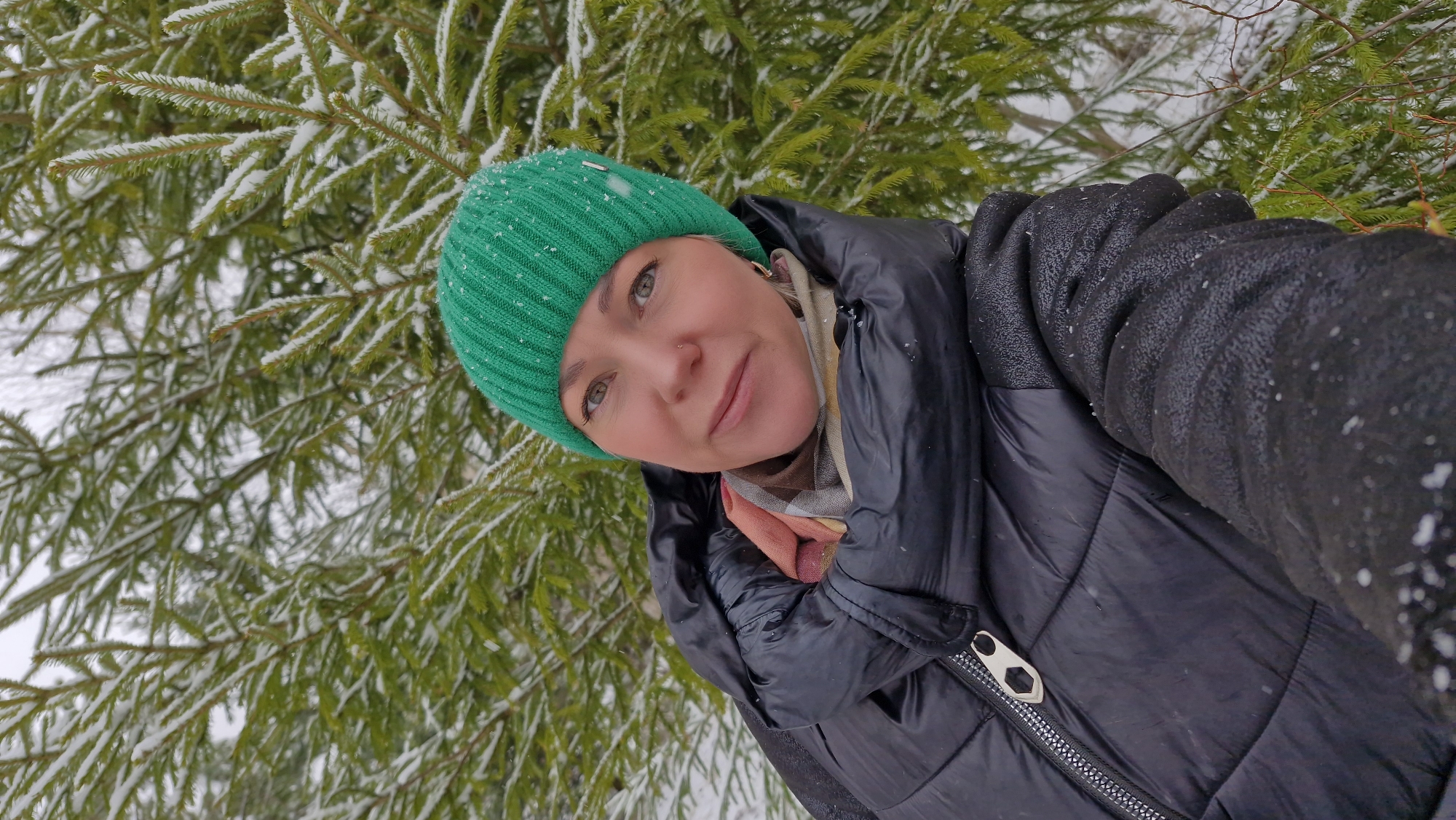 This media file features a close-up, angled selfie of a woman set against a snowy winter forest backdrop in Otchor, Russia. The woman, who is the primary subject, has light-colored eyes, a small nose piercing, and a subtle, content smile as she looks towards the camera. She is dressed in warm winter attire, including a bright green knitted beanie hat, a black puffer jacket, and a multi-toned scarf with visible shades of pink, brown, and white. Her right arm, covered by a black glove dusted with snowflakes, is visible on the right side of the frame.
The background is filled with dense evergreen trees, likely spruce or fir, their needles and branches heavily coated with fresh white snow. The ground beneath the trees is also snow-covered, visible in patches. Light snowfall is actively occurring, with numerous small snowflakes visible on her hat, jacket, and gloves, emphasizing the cold, wintry weather. The scene is illuminated by natural daylight, suggesting it was taken during the day. The overall impression is one of a serene and crisp winter landscape, with the woman seemingly enjoying the cold, snowy environment. There is no visible text in the image.