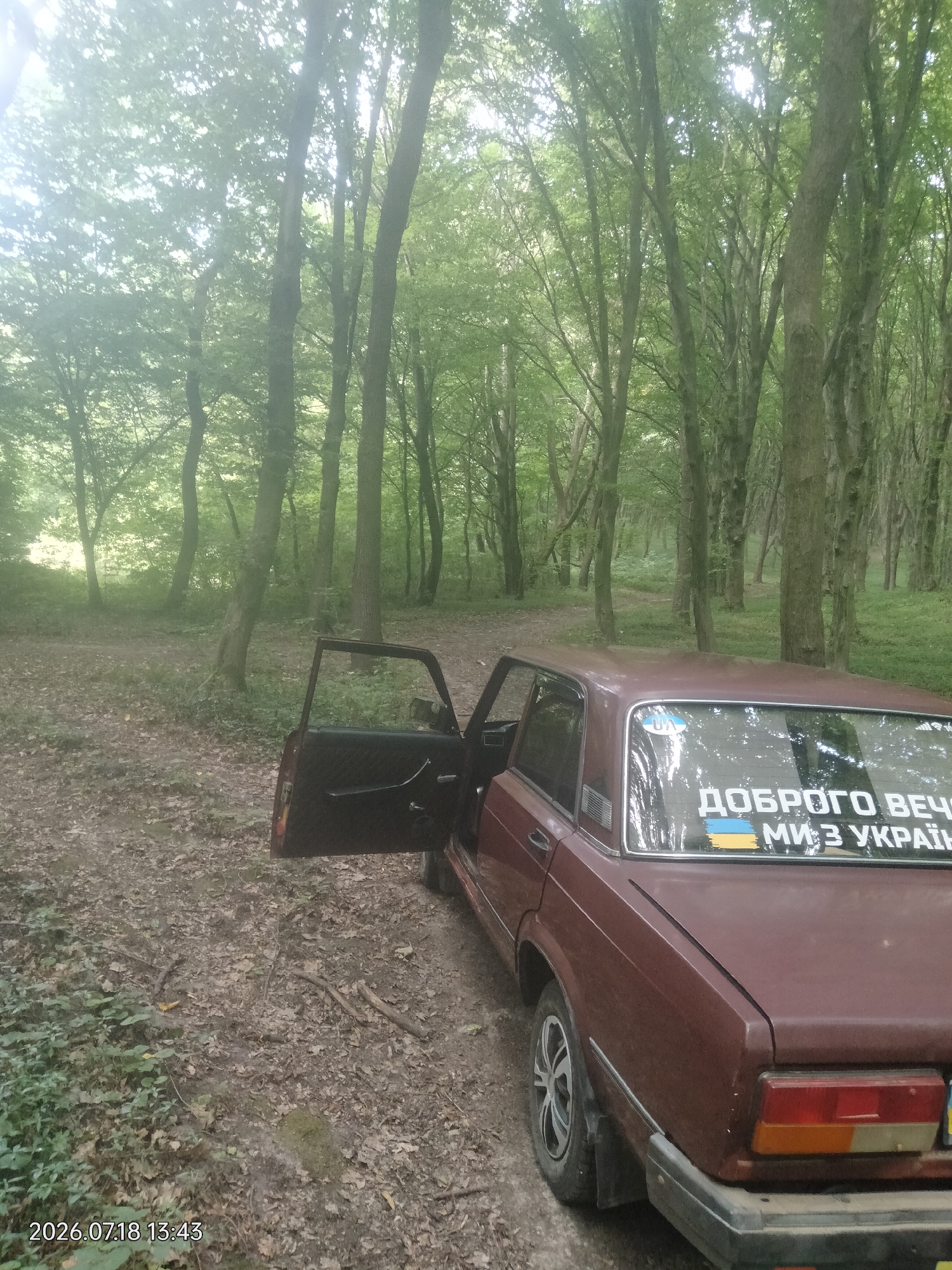A dark red, older model sedan is parked on a dirt path in a dense forest. The driver's side door is open. The car has a sticker on the rear window that reads "ДОБРОГО ВЕЧОРА МИ З УКРАЇНИ" with a Ukrainian flag emblem. The trees are tall and leafy, suggesting a spring or summer scene. The ground is covered with fallen leaves and some low-lying green foliage. The lighting is diffused, with sunlight filtering through the canopy, creating a slightly hazy atmosphere. The timestamp in the lower left corner indicates the image was taken on July 18, 2026, at 13:43.