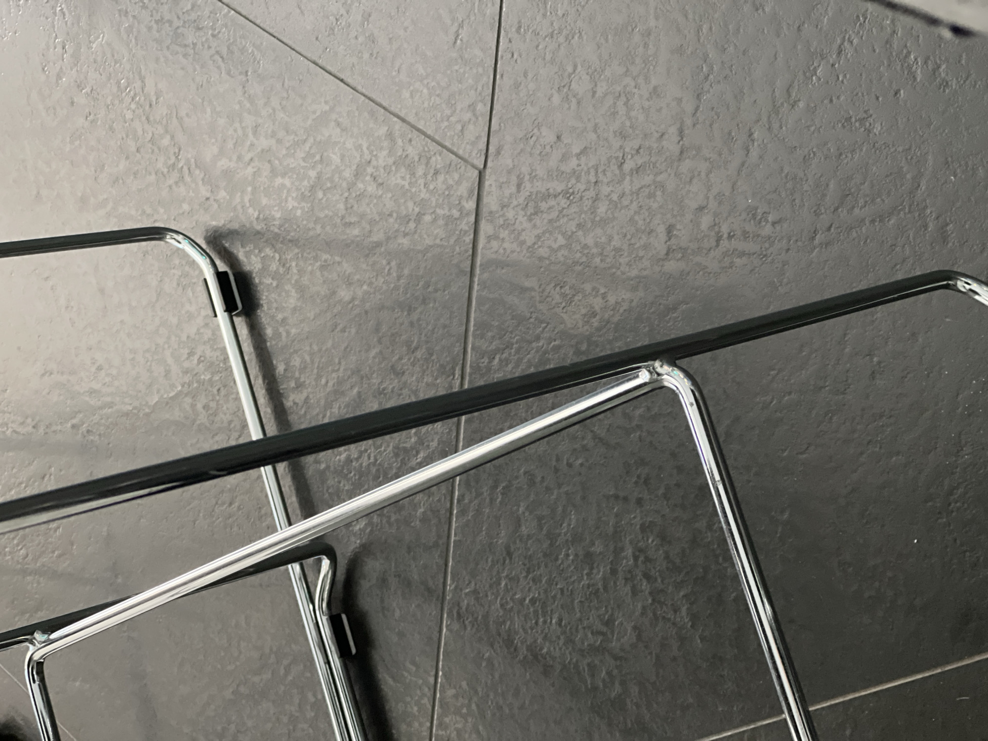 The image displays a close-up, low-angle view of the base of modern furniture situated on a tiled floor in Moscow, Russia. The primary visual subject is a structural framework constructed from highly reflective, polished chrome-plated tubular metal. Multiple segments of this tubing are visible, forming curved and straight elements of the furniture's support structure. Black, rectangular plastic or rubber glides are affixed to at least two points on the horizontal sections of the metal frame where they make contact with the floor. The flooring consists of large, dark gray to black tiles, exhibiting a textured, somewhat mottled surface. Lighter-toned grout lines are visible, defining the edges and intersections of these tiles across the floor plane. Reflections from ambient overhead lighting are evident on both the metallic surfaces and the floor. No individuals, specific actions, or other distinct objects are present in the scene.