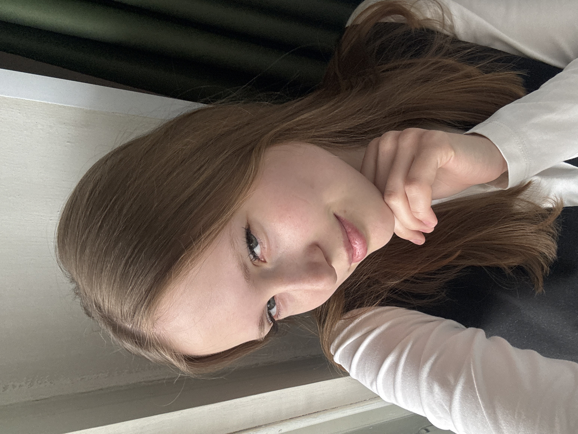 A young woman with light brown hair is positioned in front of what appears to be a window, with a section of dark green curtain visible in the background. The woman is wearing a white long-sleeved shirt over a black garment. She is resting her chin on her hand, with her fingers curved around her face. The location cannot be determined to be Chaikovsky, Russia from the image.