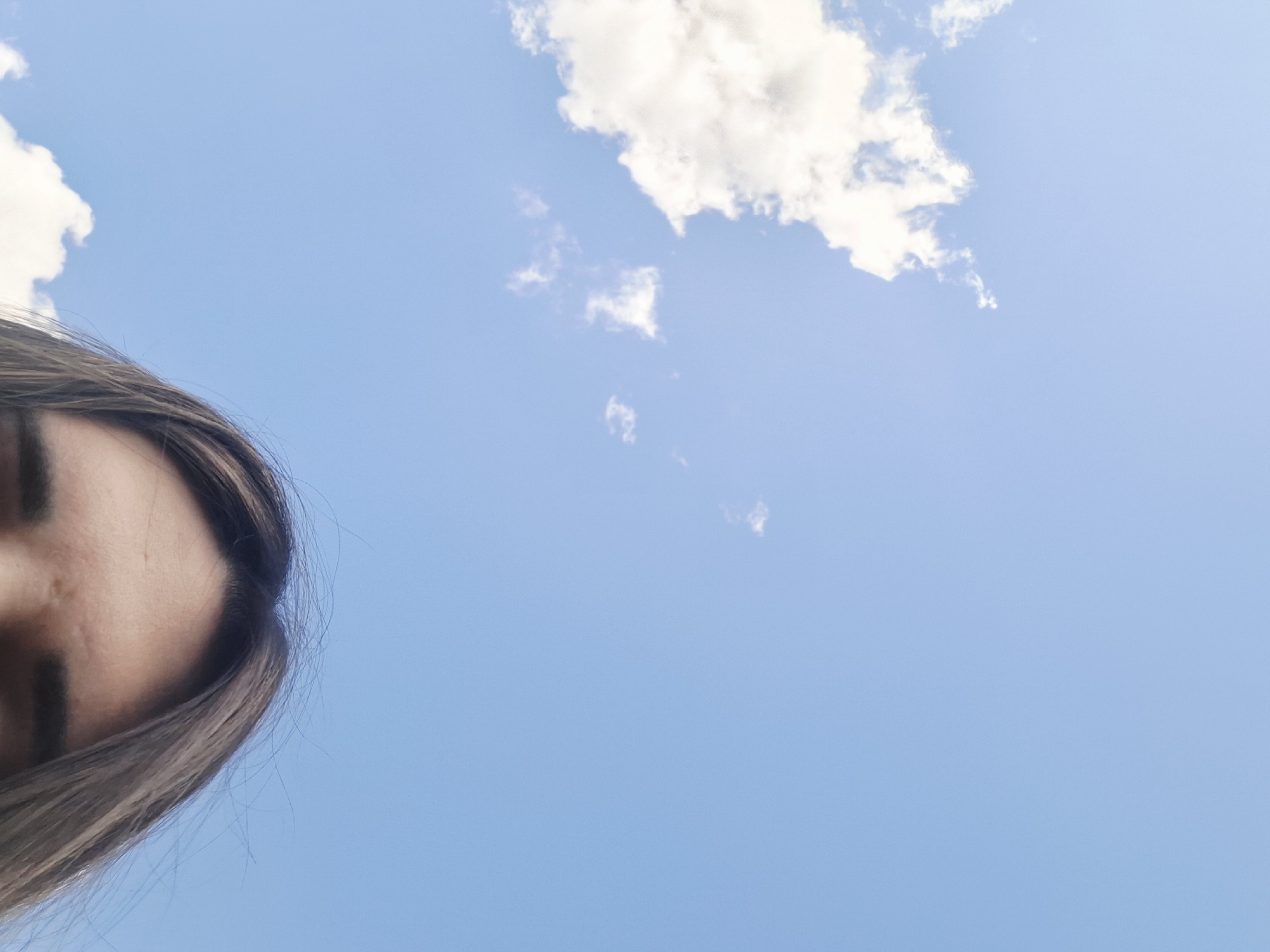 The image is a low-angle shot of a person's face and hair against a bright blue sky with white clouds. Only a portion of the person's face, including their forehead and part of their eye, is visible on the left side of the frame. Their hair, which appears to be brown and extends down the left edge of the image, is also captured. The sky is clear with scattered white, fluffy clouds, suggesting daytime. There are no discernible objects or text in the image. The overall impression is one of looking up towards the sky, perhaps outdoors on a pleasant day. The composition is minimalistic, focusing on the contrast between the sky and the human subject. The lighting indicates it is likely midday, with the sun high in the sky.