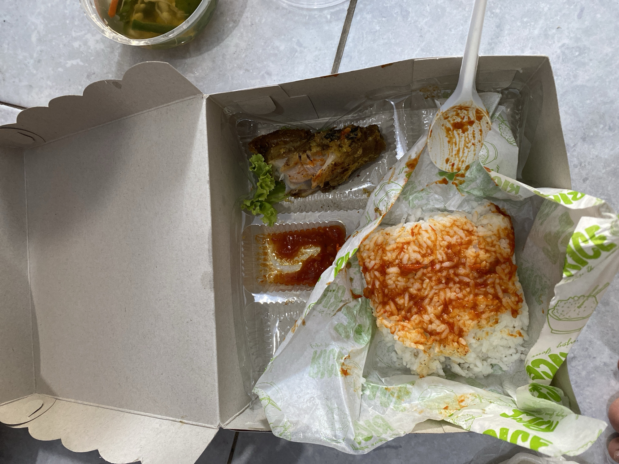 The image shows a white cardboard takeout box, open and placed on a light-colored, possibly tiled surface. Inside the box, there are three main food items: a portion of white rice topped with a red sauce, a small container of red sauce, and a piece of fried chicken with a small garnish of lettuce. A white plastic spoon with traces of sauce rests on the edge of the box, near the rice. To the right of the box, a small, clear plastic container holds pickles or pickled vegetables. The overall scene suggests a meal that has been opened for consumption. The environment appears to be indoors, with diffused lighting. There is no clear indication of the specific location within Madiun, Indonesia, beyond it being a place where such food would be served or consumed.