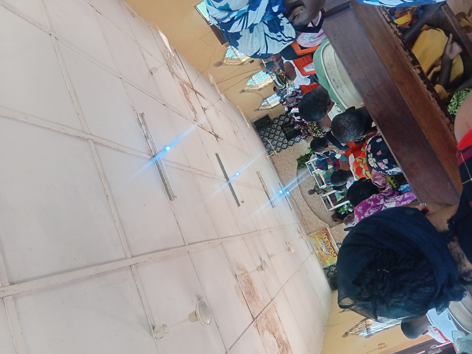 The image shows the interior of a church in Ejule, Nigeria, during a service. The ceiling is white with exposed beams and features several ceiling fans and fluorescent lights. The walls are a pale yellow, and arched windows with intricate metal grates are visible. The church is filled with people, many of whom are wearing colorful head coverings and traditional clothing. They are seated on wooden pews. In the background, a banner with text that appears to be in English is displayed. A man is standing at a pulpit area, likely leading the service. The overall scene suggests a community gathering for worship.