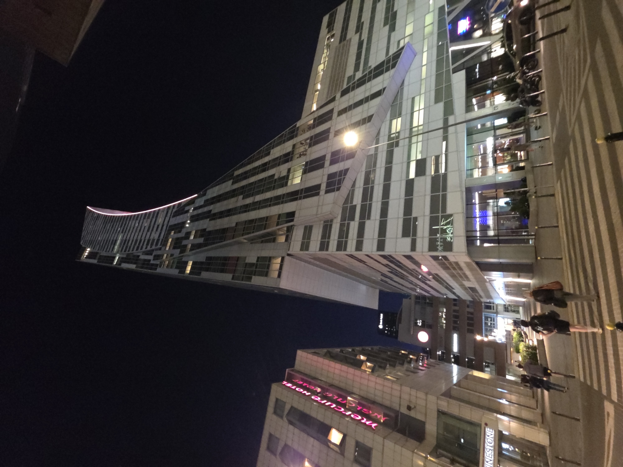 The image is a low-angle shot at night in Warsaw, Poland, focusing on modern architecture and street life. A towering, predominantly white building with a unique, curved design and diagonally striped window patterns dominates the left side of the frame. The building is illuminated, with a pink neon light tracing its curved edge. To the right, the "Mercure Hotel" is visible, identified by its magenta neon signage. People are walking on the brightly lit sidewalk. The sky above is dark. A modern lamp post provides light to the scene.