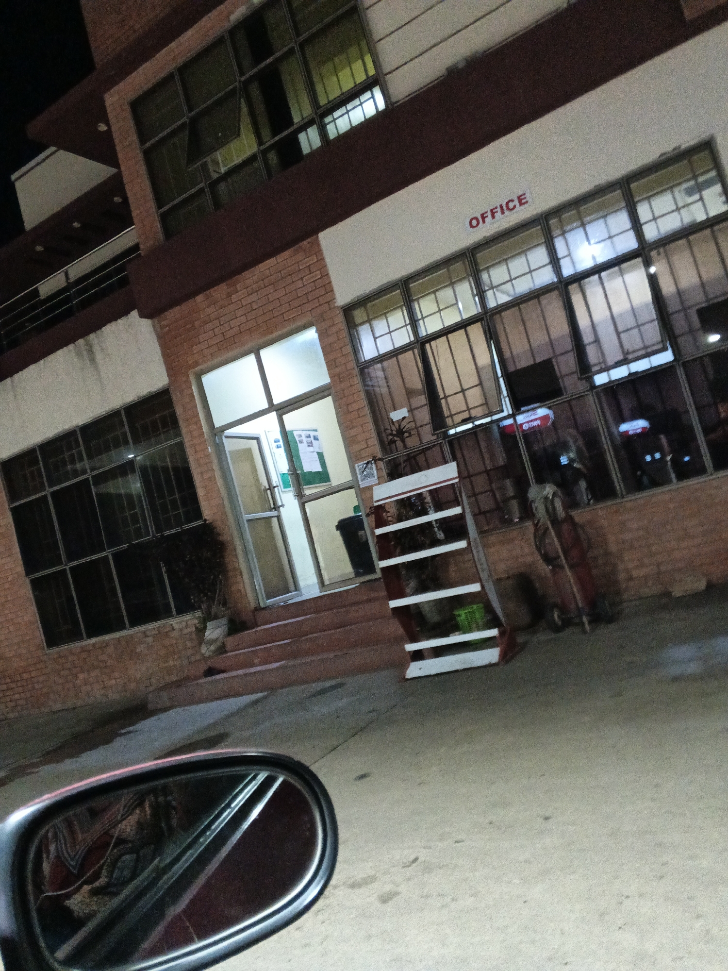 The image depicts a building with red brick facade and maroon trim, identified as an "OFFICE" with red lettering. The building has multiple large windows, some of which are open. In the foreground, a car's side mirror is visible, reflecting a pattern likely from a seat cover. The scene appears to be an exterior shot, possibly at dusk or night, given the dark sky in the upper left corner. An entrance to the building features double doors and a small staircase leading up to it. Inside the entrance, a bulletin board with papers is visible, along with a trash can. To the right of the entrance, a display rack with shelves holds a green basket and other items. Further to the right, a cart with a red handle and wheels is present. The overall environment suggests a commercial or institutional setting in Jos, Nigeria.