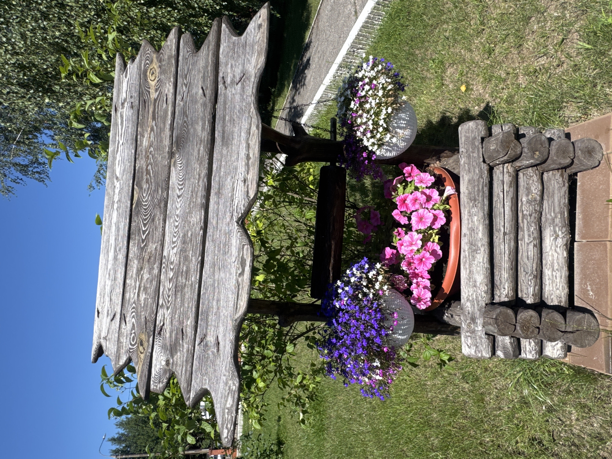 A decorative wooden well structure is positioned outdoors on a sunny day. The well is adorned with several potted plants and hanging baskets filled with colorful flowers. The structure itself is made of weathered wood, with a multi-layered roof and a base constructed from logs. In the background, green grass, trees, and a paved walkway are visible, all under a clear blue sky. The scene suggests a garden or park setting.