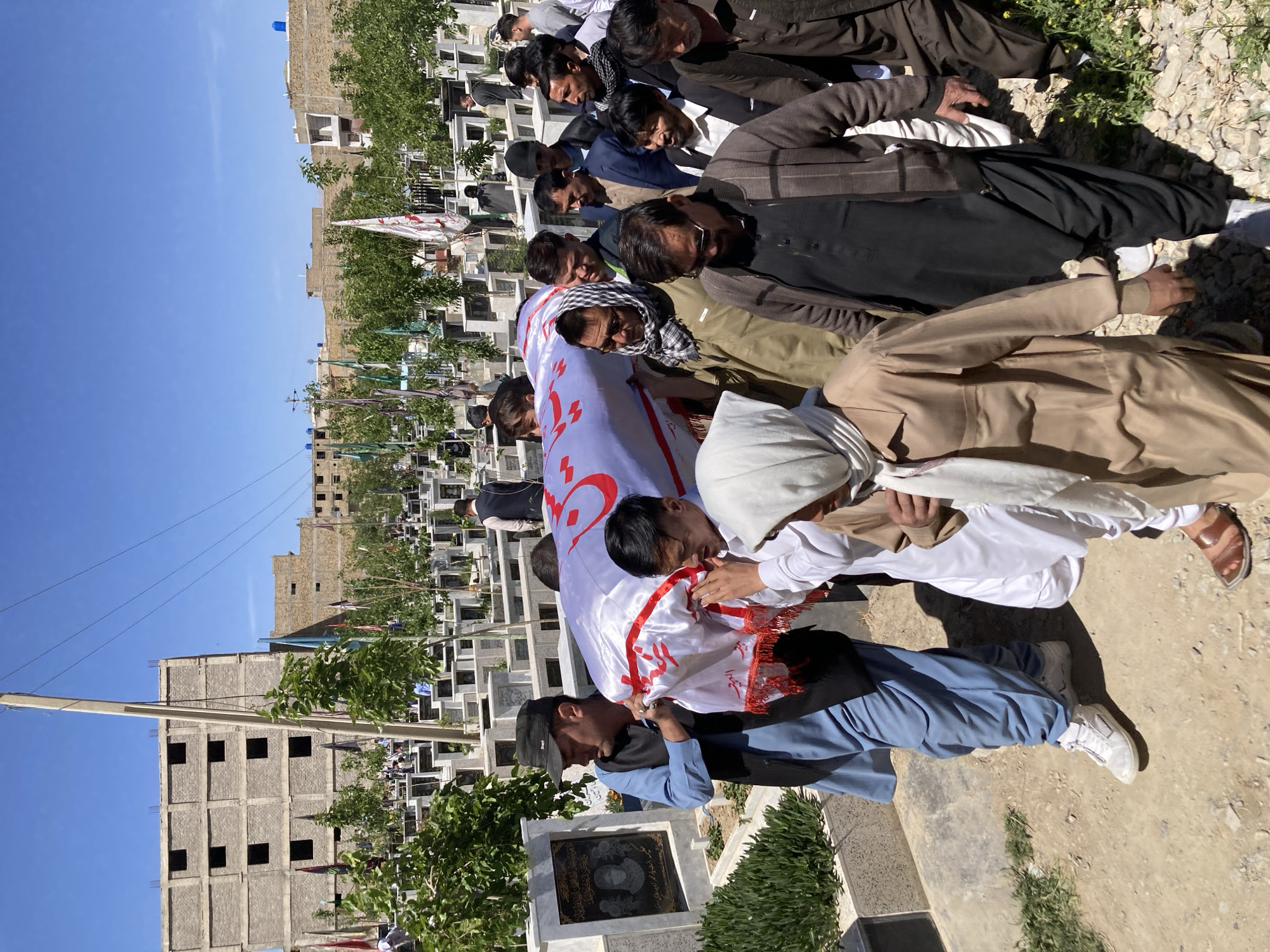 The image shows a funeral procession taking place in what appears to be a cemetery, in Quetta City Tehsil, Pakistan. A group of men are carrying a coffin draped in a white cloth with red writing. The mourners wear a variety of clothing, some in traditional Pakistani dress and others in more Western attire. The cemetery is crowded with graves and headstones, and in the background, there are buildings, including an unfinished building under construction. The sky is blue and the sun is shining. The general mood is somber and respectful, given the context of a funeral.