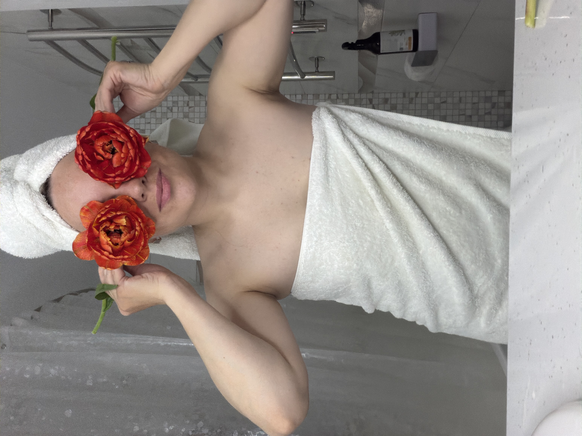 A vertically oriented photograph captures an individual in a modern bathroom setting. The subject, an individual with fair skin, is positioned horizontally across the frame, suggesting they are lying down or the image has been rotated 90 degrees clockwise. They are wrapped in a white body towel from the chest down, and a second white towel is wrapped around their head like a turban.
The person holds two vibrant red and orange flowers, resembling parrot tulips, over their eyes, one in each hand. A subtle smile is visible on their lips, conveying a relaxed and playful expression. Their arms are gently raised, supporting the flowers in place.
The background, consistent with a bathroom in Votkinsk, Russia, features light-colored tiled walls and a chrome towel rack behind the person's head. Further back, a section of wall with mosaic-style tiles is visible, along with a wall-mounted dispenser or bottle, which has an unreadable label. To the right side of the frame, a clear glass shower screen, covered in numerous water droplets, suggests a recent shower or bath, reinforcing the serene, post-bathing atmosphere of the scene. The overall mood is one of relaxation and lightheartedness.