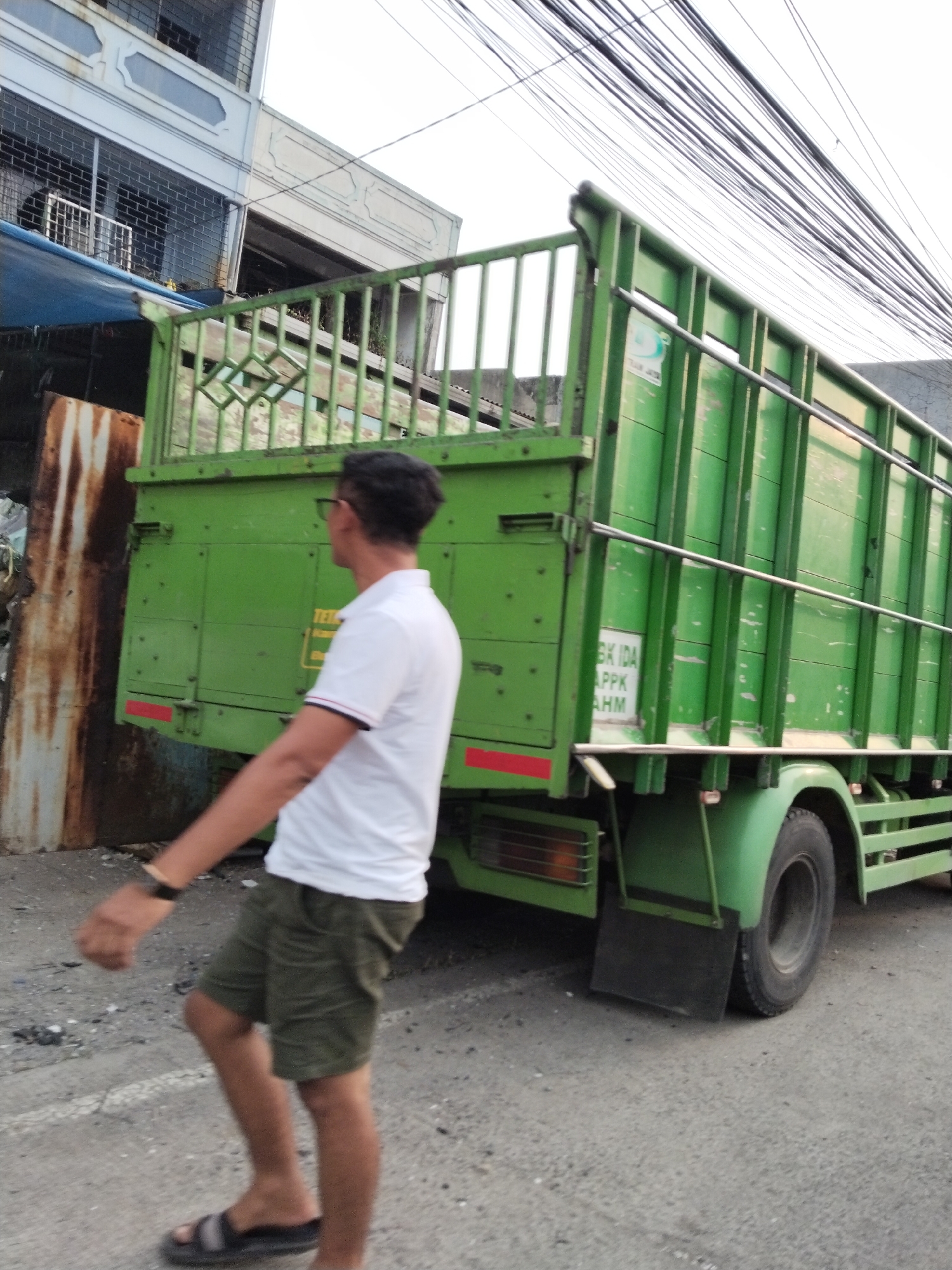 A full-shot, eye-level, outdoor, slightly blurry, vertical-orientation photo captures a man walking next to a bright green truck with a metal cage on the back. The man, wearing glasses, a white polo shirt with red and blue trim on the collar and sleeves, and olive-green shorts, is walking away from the camera. He has on black sandals. The truck, a common cargo vehicle, is parked on a paved road, and the man appears to be moving alongside it. The scene is set against a backdrop of a two-story building with barred windows on the upper level and a blue tarp covering the lower section. Overhead, a dense network of black electrical wires stretches across the sky, indicating an urban environment. The lighting suggests it is either early morning or late afternoon. There is visible text on a white sticker on the truck's side, appearing to be in an Indonesian script. The overall atmosphere is that of a casual street scene in an Indonesian city.