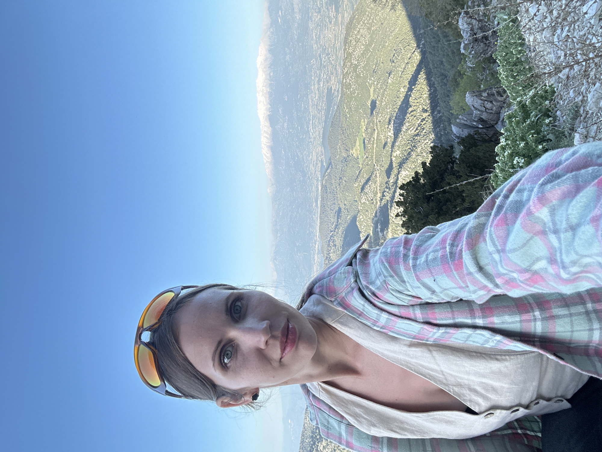 A woman with light skin and brown hair pulled back by sunglasses is taking a selfie outdoors. She is wearing a light pink and green plaid shirt over a light-colored button-up shirt. She is standing on a rocky, mountainous terrain with sparse vegetation. In the background, a vast landscape stretches out, including distant mountains and a city. This location is likely on a high point overlooking the city of Fethiye, Turkey, given the described environment and topography. The weather appears clear and sunny.