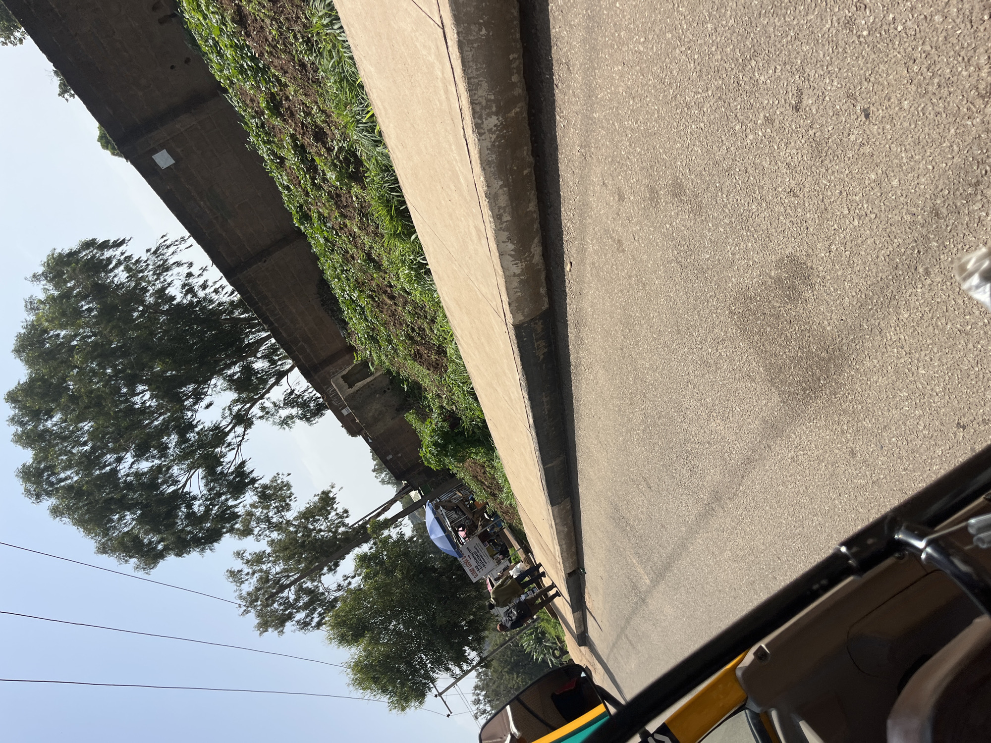 The image captures a street scene in Bukuru, Nigeria, on a bright, sunny day. A paved road occupies the foreground, with a concrete sidewalk bordering it. Beyond the sidewalk, a vegetated embankment rises, topped by a brick wall. Several trees with lush green foliage are visible, some reaching towards the clear blue sky.
In the middle ground, a few individuals are seen standing on the sidewalk near a small stall covered by a blue umbrella. A sign is mounted on a pole behind the stall. A three-wheeled vehicle, possibly a rickshaw or auto-rickshaw, is partially visible in the lower left quadrant of the image, suggesting the perspective is from within or alongside such a vehicle. The environment appears to be a somewhat urban setting, with evidence of both natural elements and man-made structures.