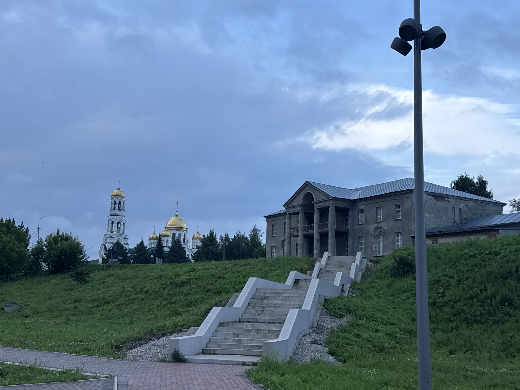 The image shows a daytime view of an exterior scene in the city of Lysva, Russia. In the foreground, a red brick walkway leads up a grassy hill. A concrete staircase with multiple landings and guardrails ascends the hill. On the right side of the frame, a tall, gray lamppost with three lamps is visible.
Dominating the midground is a light gray, two-story building with a portico supported by columns. The building appears weathered and has multiple windows. Behind this building and to the left, a white bell tower and several gold-domed orthodox churches are prominent. The churches and bell tower are located on a higher elevation.
The environment is characterized by a cloudy, overcast sky. Green grass covers the slopes of the hill, with some trees and foliage visible in the background. No people are present in the image. The scene is static and depicts a view of a historical or religious complex in an urban setting.