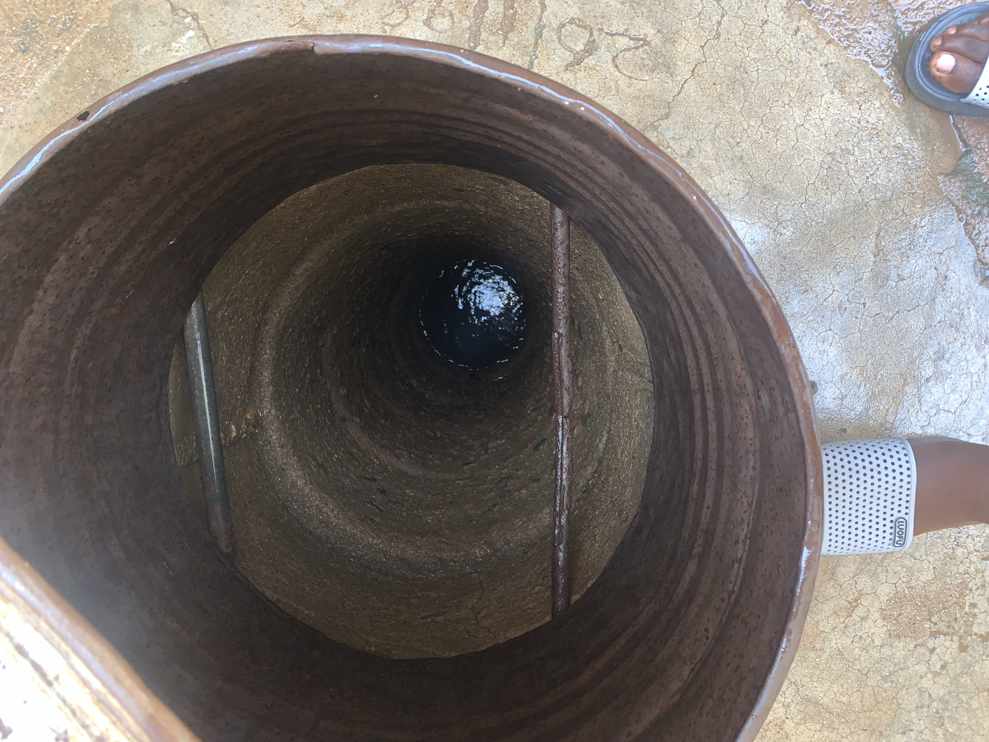 An overhead photograph depicts a deep, cylindrical well or borehole in Rafin Par, Nigeria. The well's opening is lined with a heavily rusted, corrugated metal casing, exhibiting shades of dark brown and reddish-brown. The interior of the well descends, revealing a rough, textured lining that appears to be made of concrete or stone. Approximately one-third of the way down the visible shaft, a light grey or metallic bar spans the diameter. Further below, a second, heavily rusted, brown bar is horizontally positioned. At the deepest visible point of the well, dark water is present, reflecting a bright, irregular light source from above.
In the foreground, a person's lower legs and feet are positioned over the well's edge. The person is wearing white, open-toed sandals featuring a perforated pattern. The left sandal clearly displays the brand name "WOFU" in white lettering on a black oval, oriented upside down relative to the viewer. The ground surrounding the well is a light-colored, cracked concrete or paved surface, showing areas of dampness and residual water. The visible skin on the feet is dark.