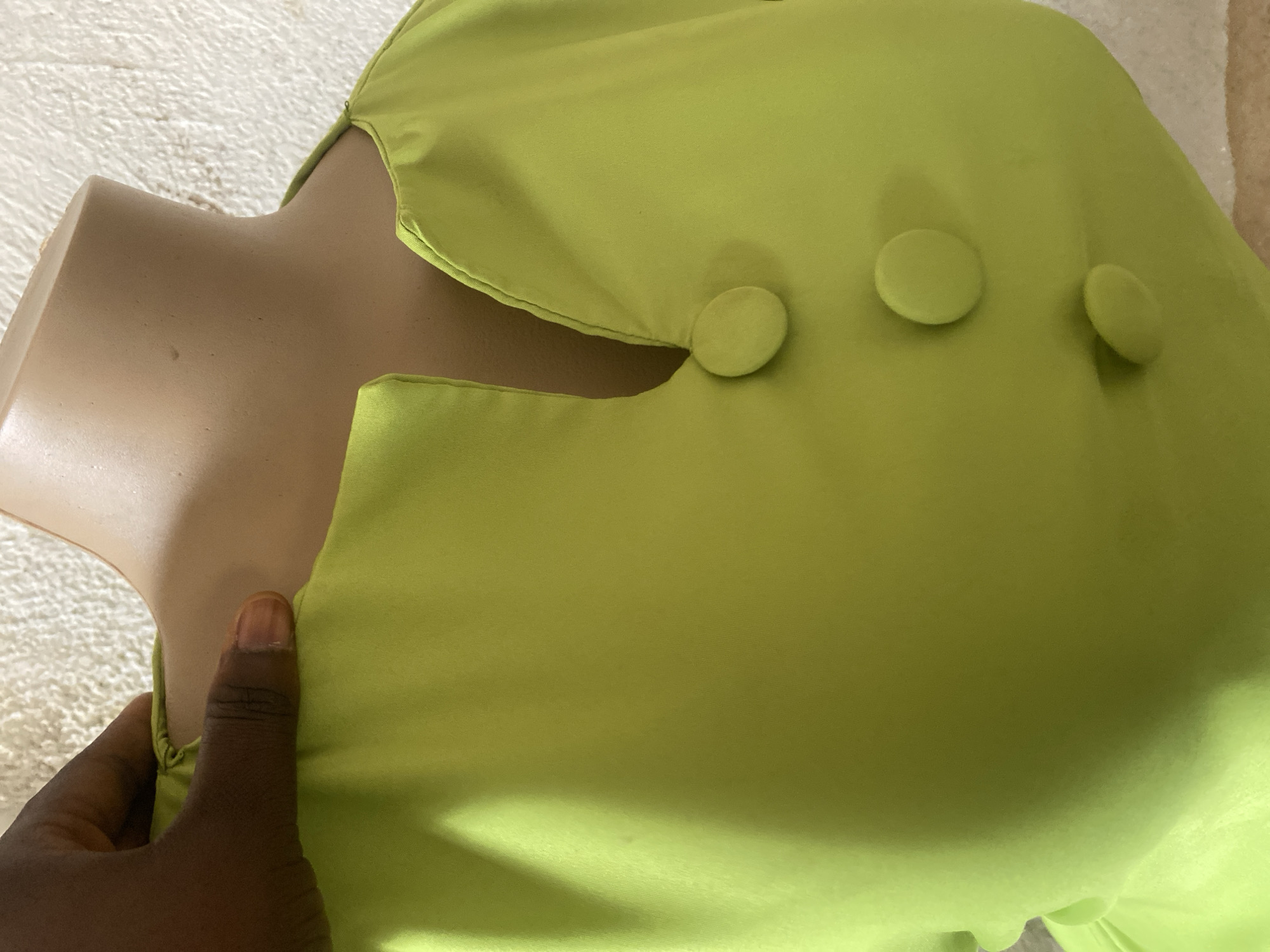 The image shows a close-up of a light green garment on a mannequin. A dark-skinned hand is holding the fabric near the neckline. The garment features three circular, fabric-covered buttons arranged vertically on the right side. The background is a textured, light-colored surface, possibly a rug or tablecloth. The lighting suggests an indoor setting. There is no visible text. The image is a detail shot, focusing on the texture and design elements of the garment. The setting and activity are not fully discernible, but it appears to be a product photograph or a detail shot of clothing.