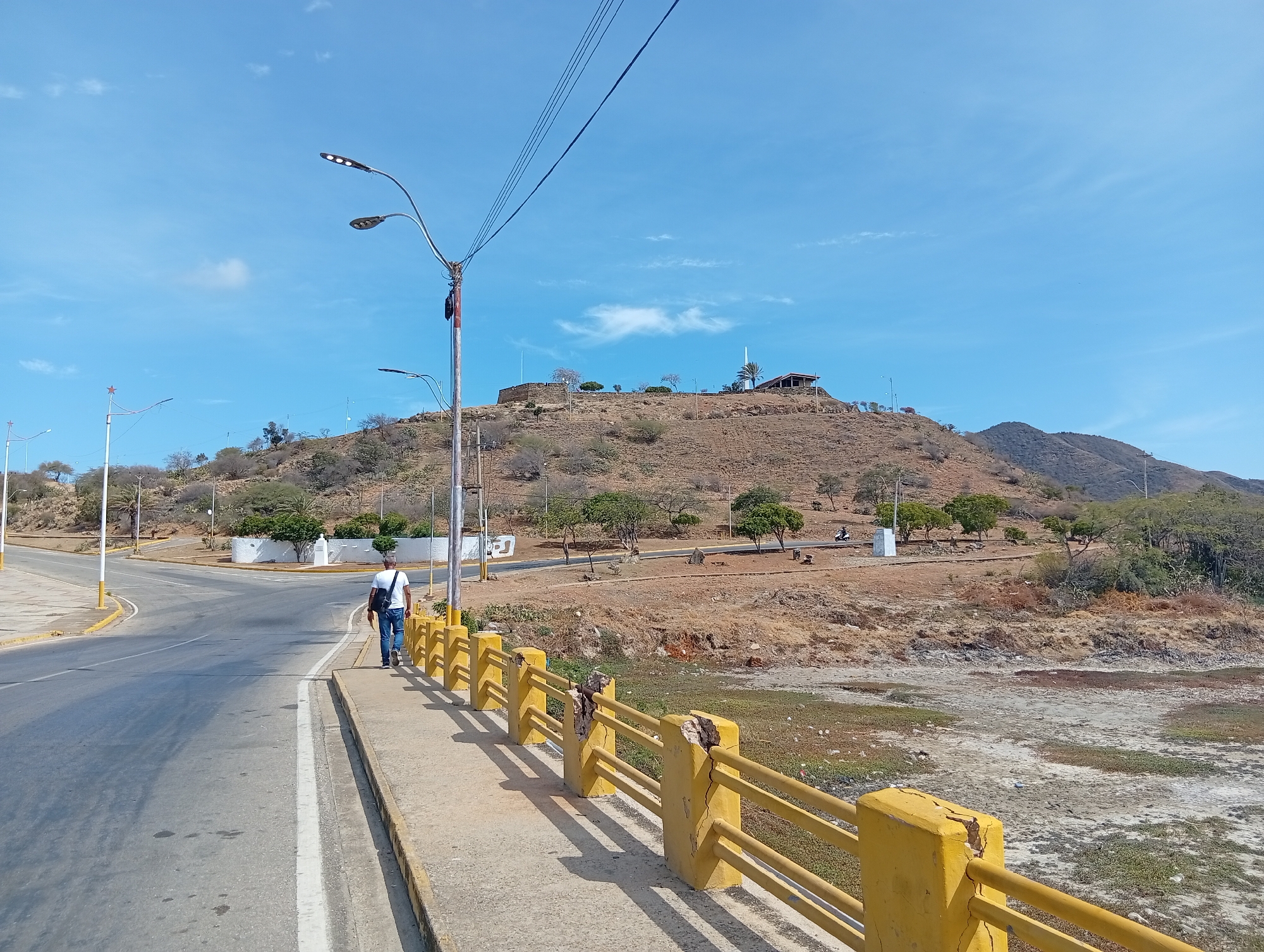 The image shows a man walking on a sidewalk along a yellow railing, next to a road on the outskirts of Juan Griego, Venezuela. The background features a large hill with sparse vegetation and structures at the summit, including a building with a tower and a stone wall structure. Power lines and streetlights are visible in the sky. The environment appears dry and arid, with dry vegetation visible around the base of the hill.