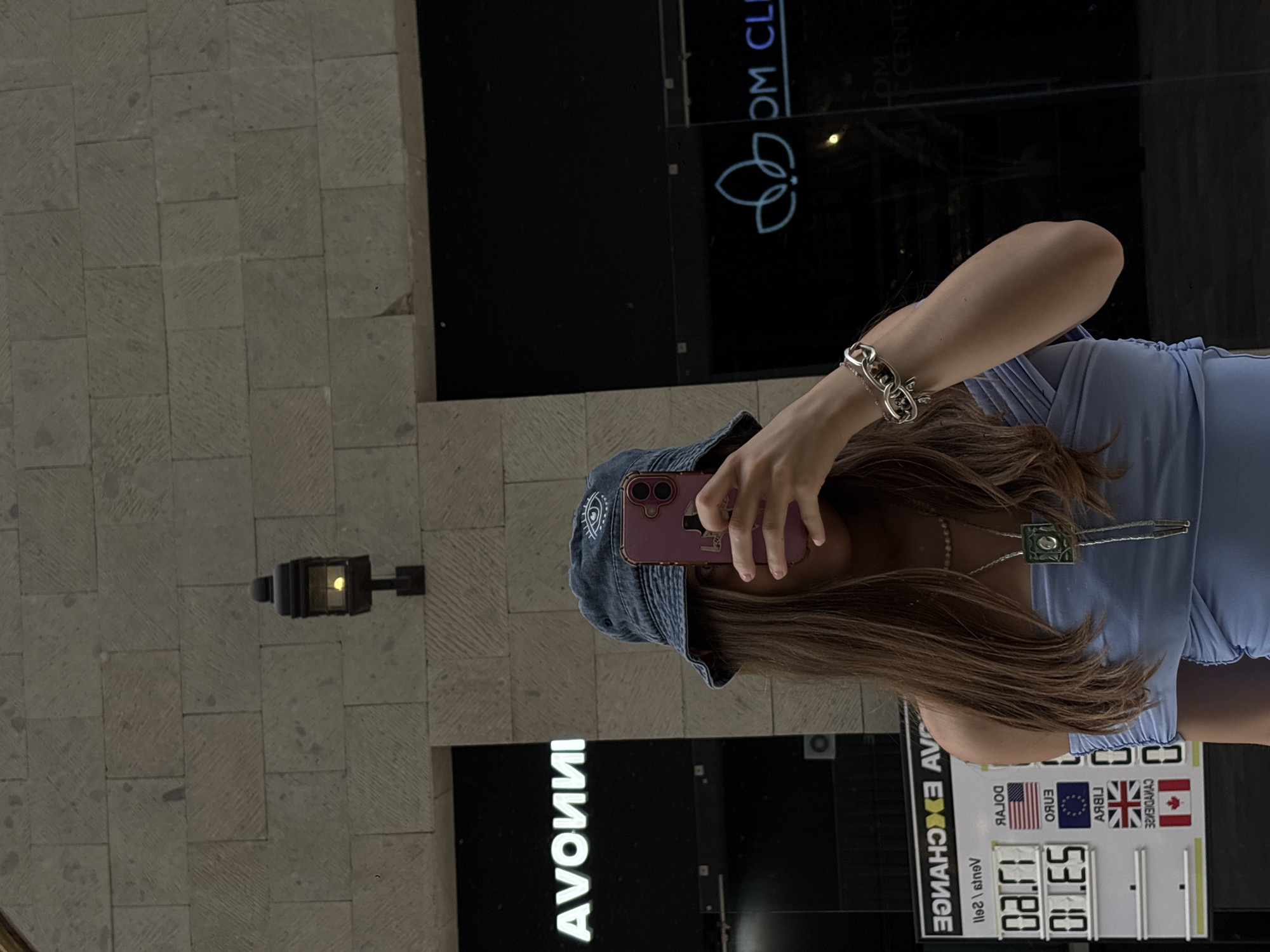 A young woman is taking a selfie in a storefront window reflection in Ciudad López Mateos, Mexico. She is wearing a blue denim bucket hat, a blue top, a silver bracelet, and a bolo tie-style necklace. She is holding a pink smartphone. Behind her, the reflection shows an Avon store and an exchange rate board displaying rates for USD, EUR, GBP, and CAD. A wall with a mounted light fixture is also visible in the reflection.