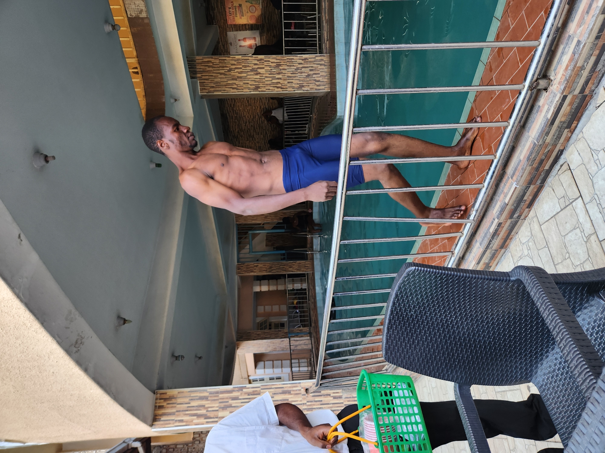 A man in blue swimming trunks stands next to a pool, leaning on a metal railing. The pool is partially visible, with turquoise water and red tiles lining the edge. The surrounding area has tiled walls and ceilings. A green plastic basket is near the edge of the scene. A person is seated in a black plastic chair near the basket. The location is Unknown, Unknown.